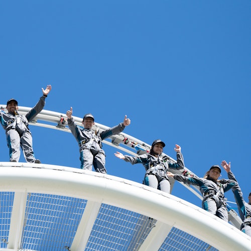 Four people in harnesses wave and smile while standing on a curved structure against a clear blue sky.