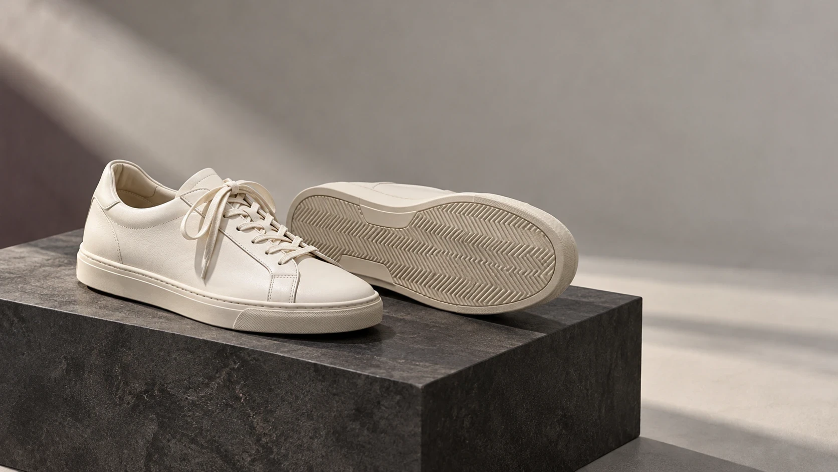 Conceptual ivory sneakers on a charcoal plinth, with one shoe showing its outsole