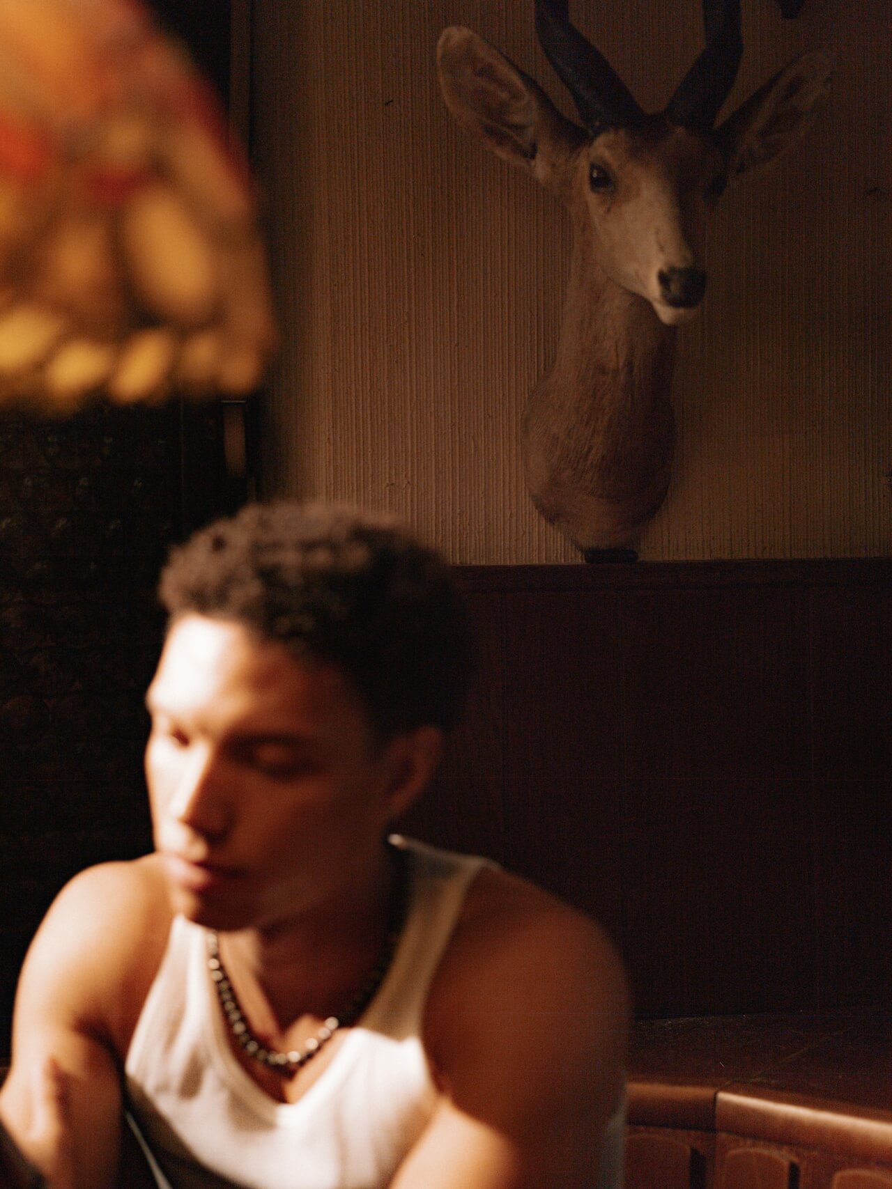 A young man in a white tank top sits pensively in dim light. A mounted deer head and an ornate lamp create a somber, reflective atmosphere.