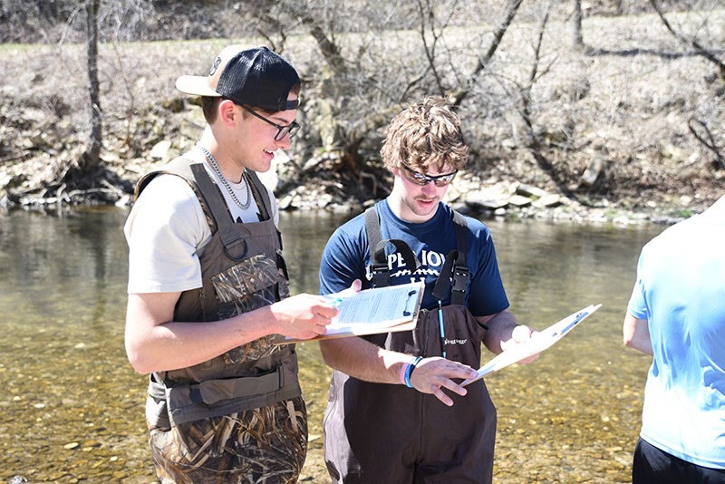 Two students wade in a river and check data on clipboards