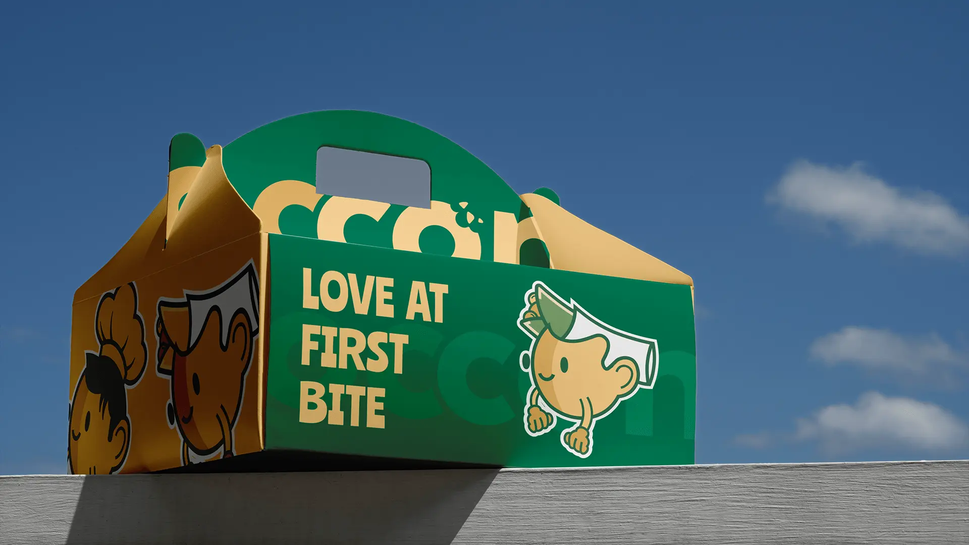 Boccone Café branded takeaway box in green and yellow with playful mascot illustrations and the slogan ‘Love at First Bite’ against a clear blue sky.