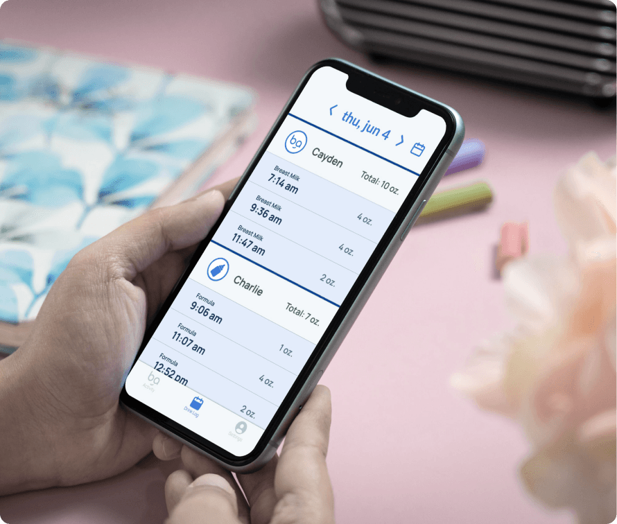 Person holding a smartphone displaying a feeding log on an app, showing detailed entries for tracking baby feeding times and amounts.