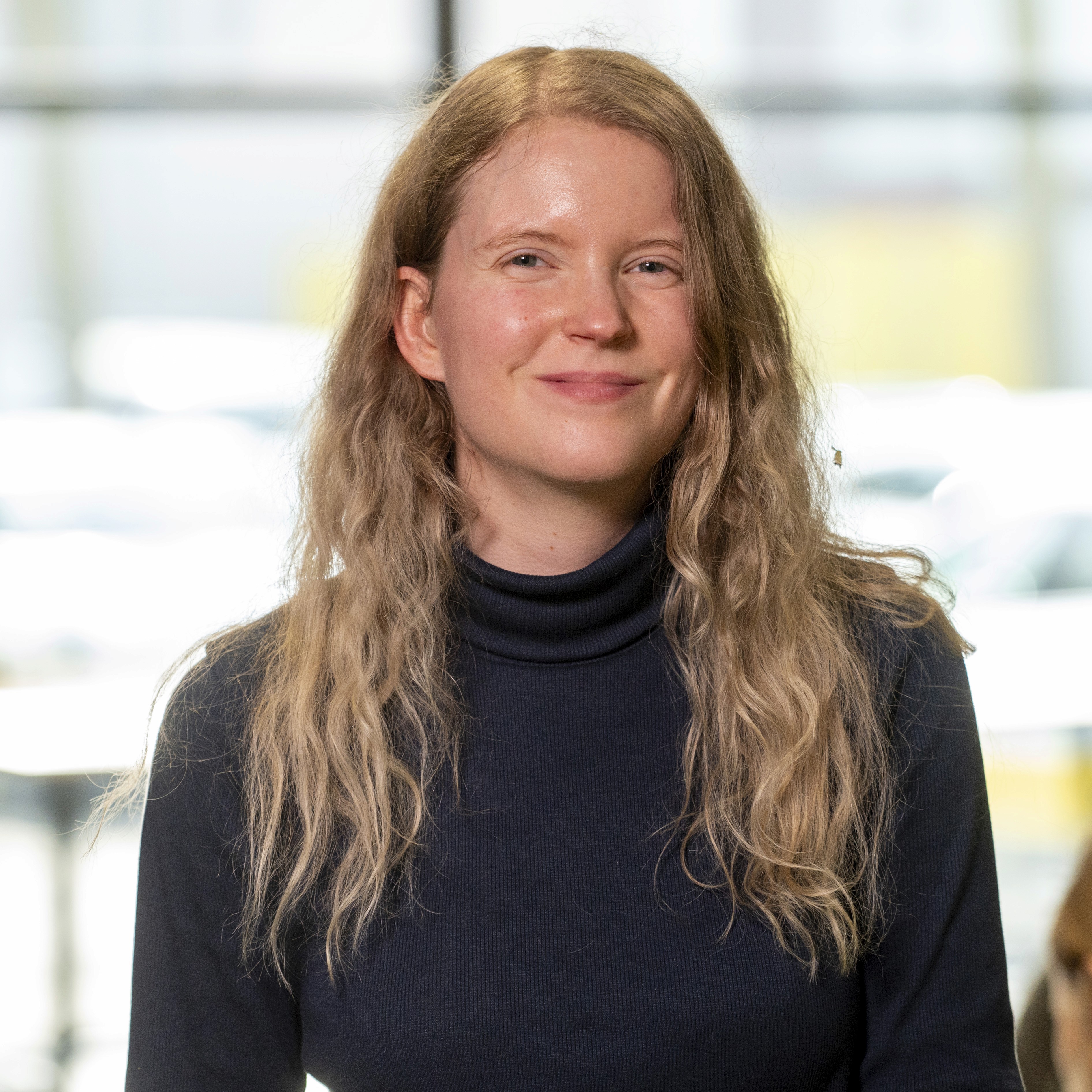 A woman with red hair and a black sleeveless top smiling confidently in a bright, modern indoor setting.