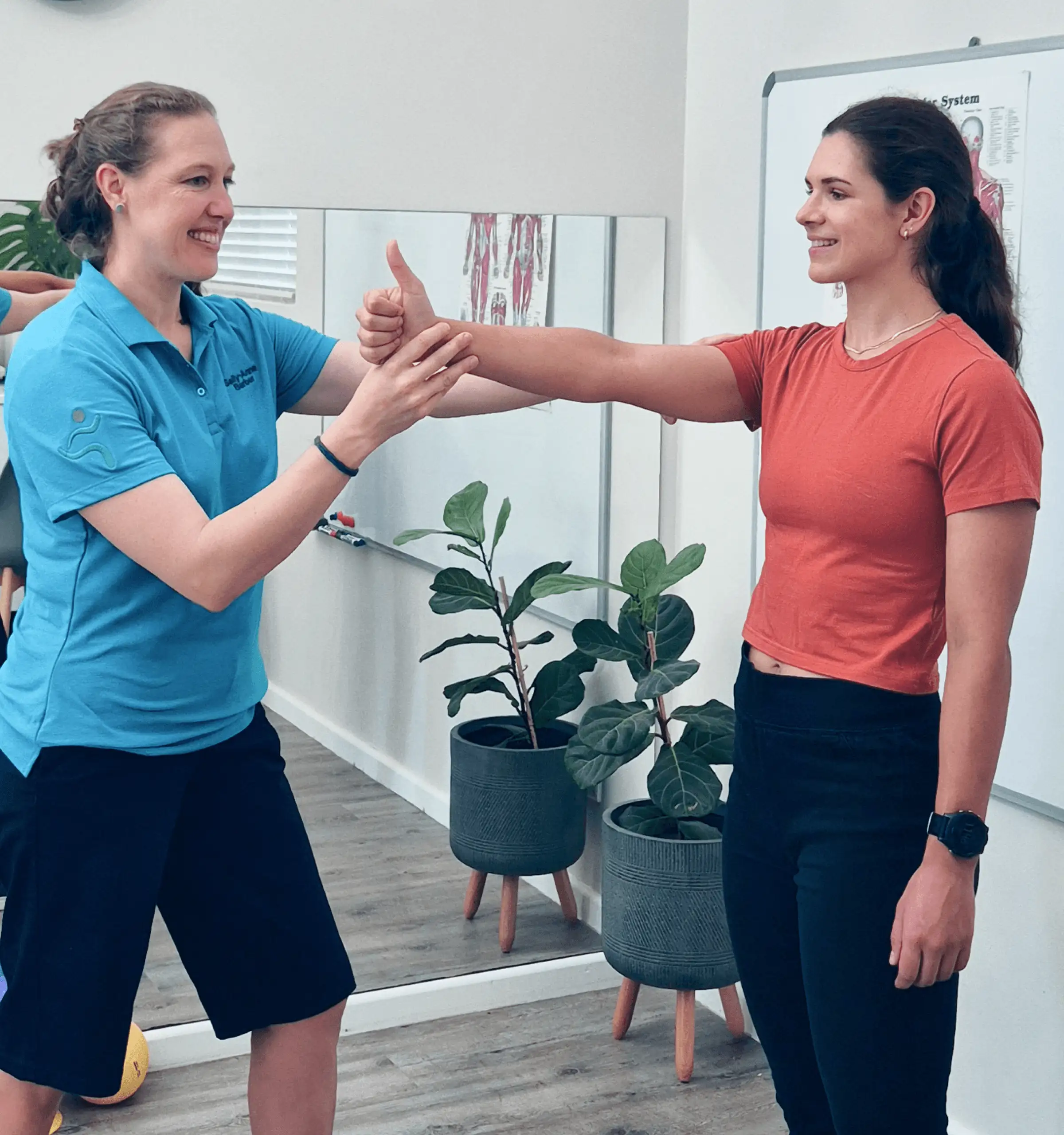 A fitness trainer assists a client by demonstrating an exercise in a bright, plant-filled indoor space.