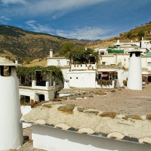 白墙平顶的房屋和烟囱,坐落在山坡上,背景是连绵的群山和蓝天。