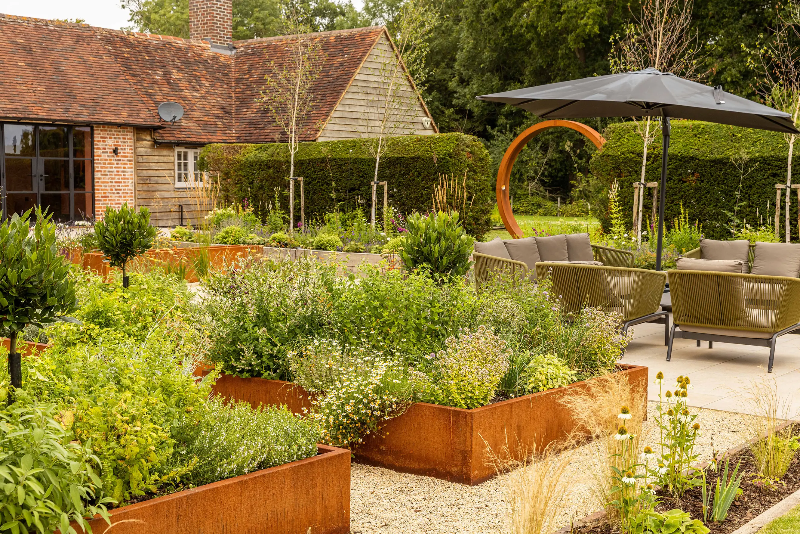 A lush garden featuring raised herb beds, seating areas, and a patio umbrella surrounded by greenery.