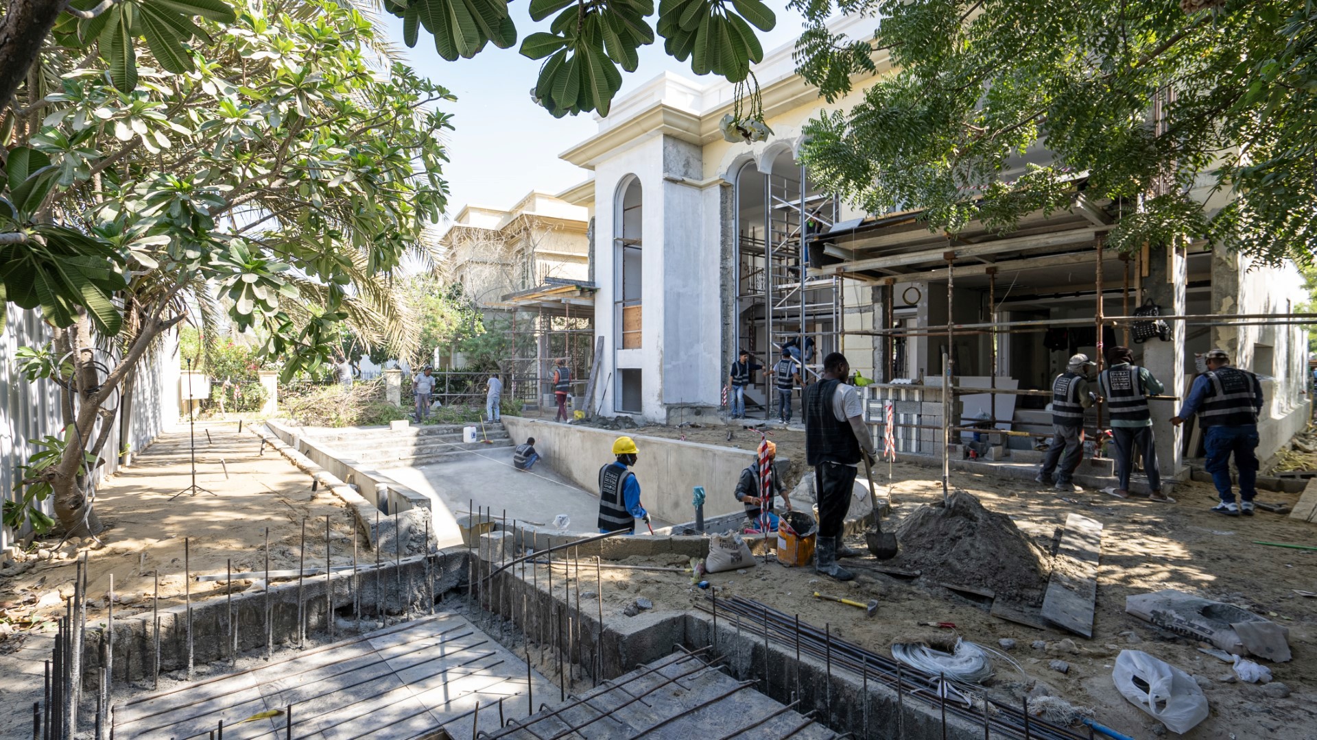A construction site in Jumeirah Islands with multiple workers in hard hats operates around a large residential building under renovation, featuring scaffolding, newly installed windows, and concrete walkways, with lush green trees in the background.
