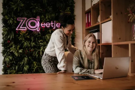 A person stands behind a woman at a desk, both engaged with a laptop in a stylish office space.