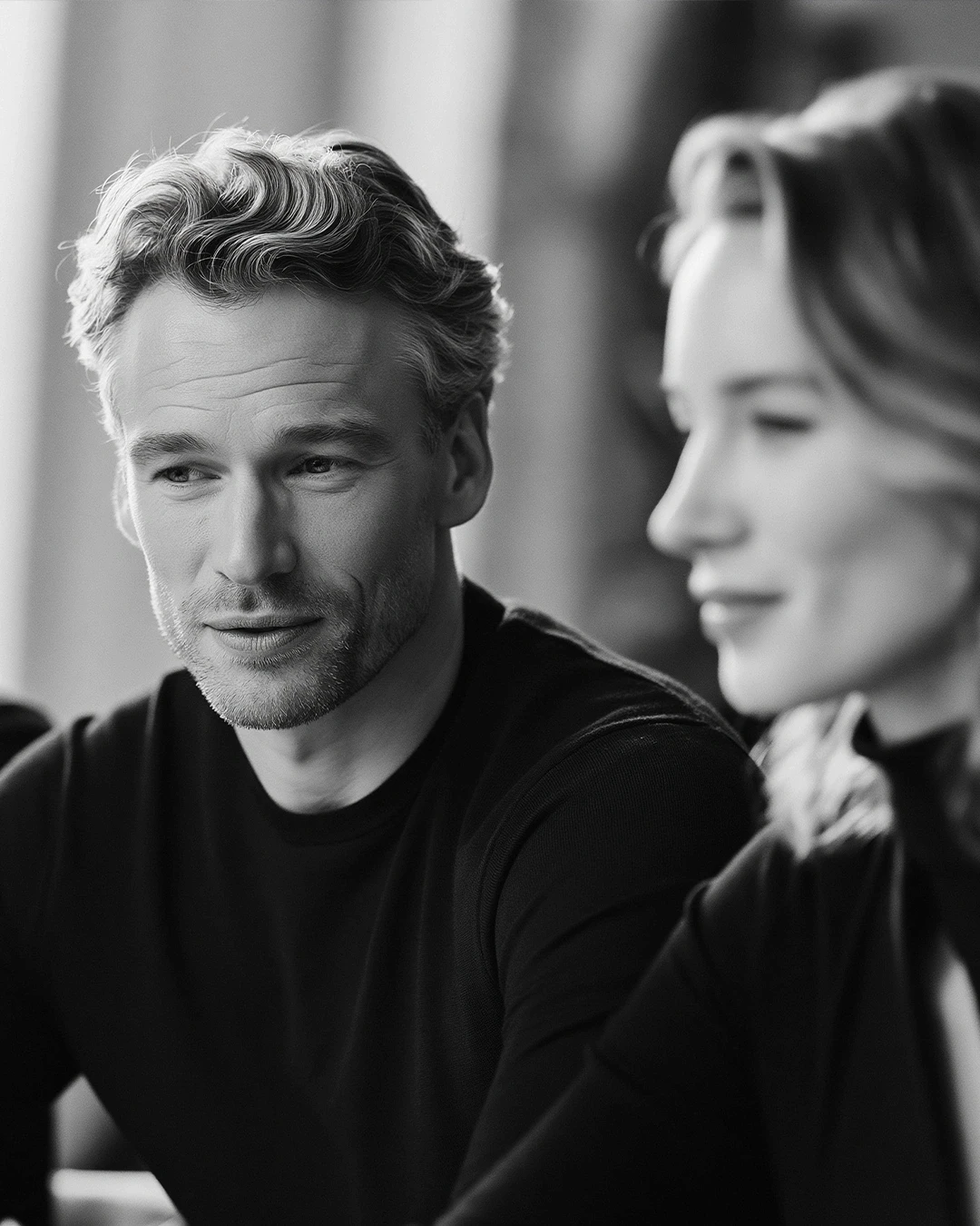 A black and white portrait of a man with wavy hair looking thoughtfully to the side, with a blurred woman in the foreground.