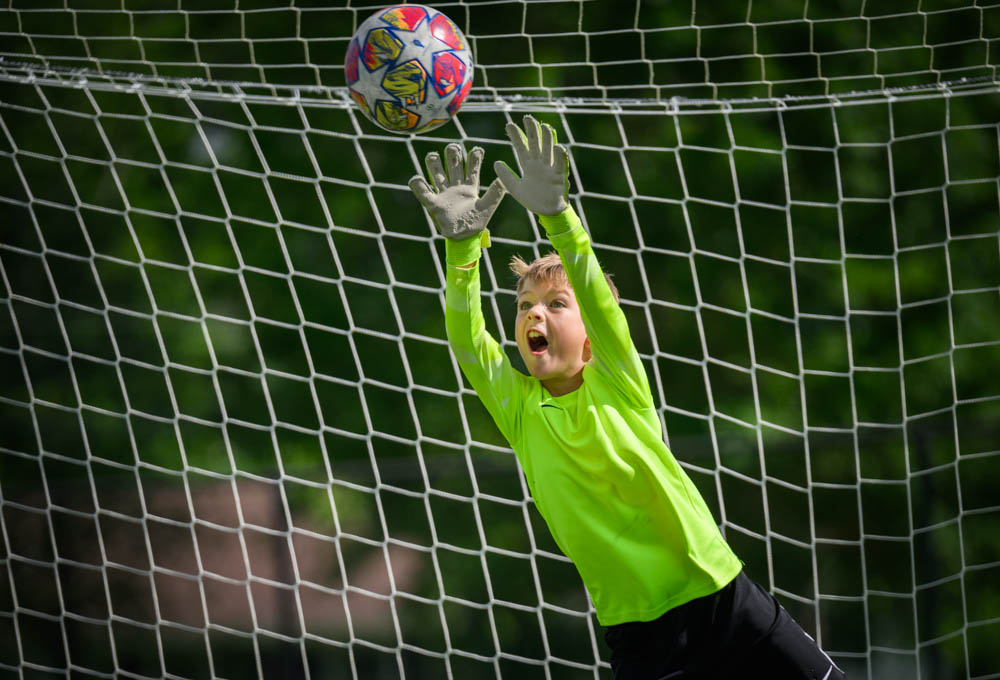 A youth goalkeeper in a bright green jersey jumps with both arms raised, mouth open as he shouts while making a save.
