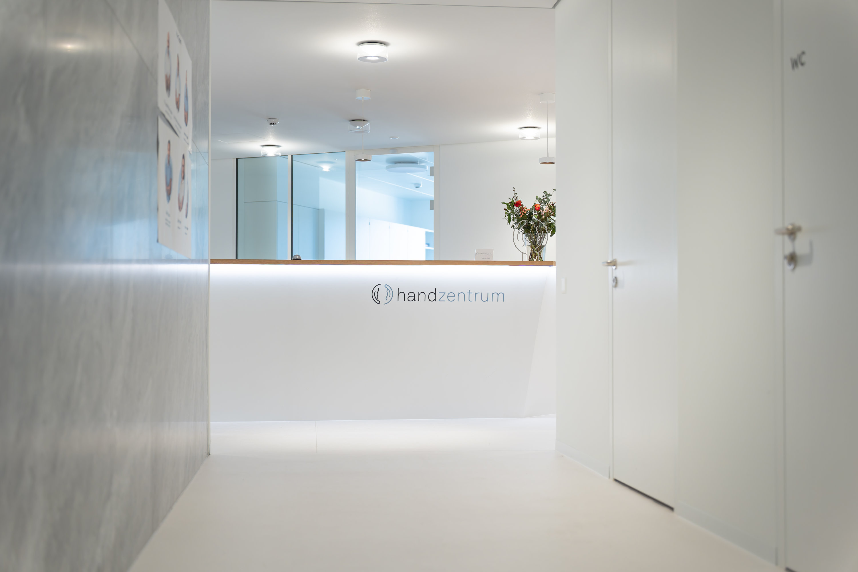Ein moderner, minimalistischer medizinischer Empfangsbereich mit einer weissen Theke mit der Aufschrift Handzentrum Pratteln, einer Vase mit Blumen, heller Beleuchtung und weissen Wänden.