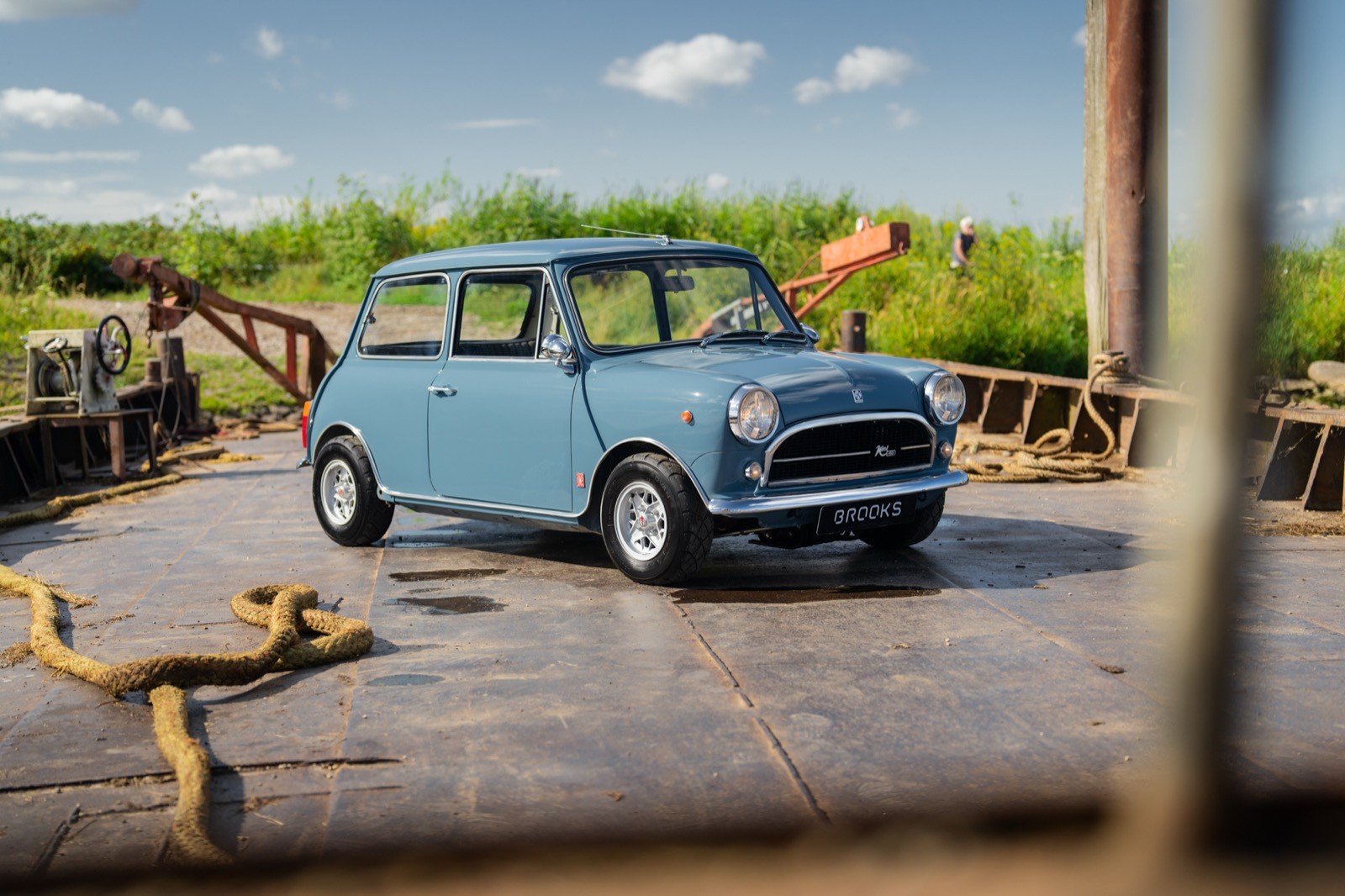 A vintage blue car parked in an open area with green surroundings and a clear blue sky.