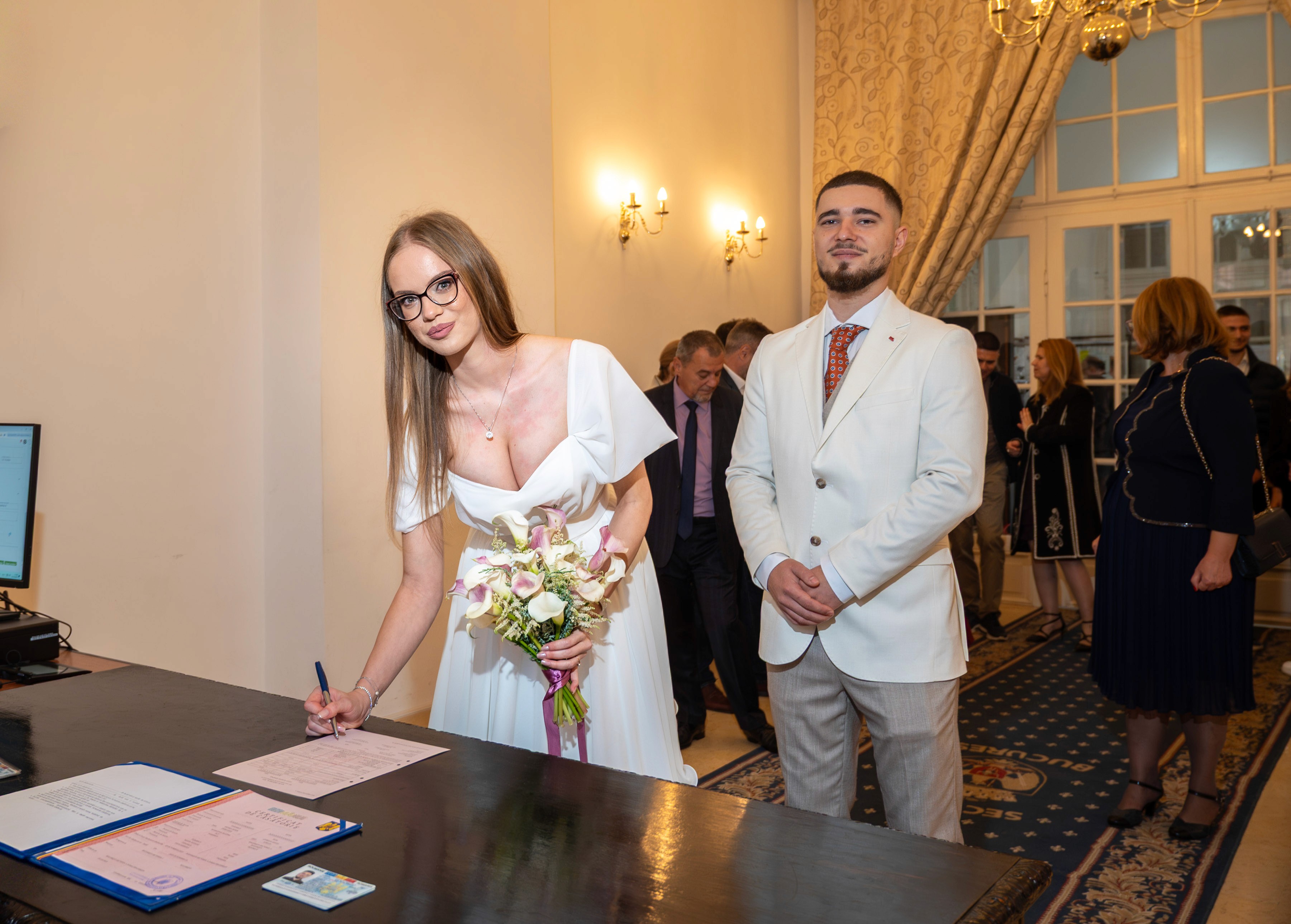 Soție semnând actele la cununia civilă, în timp ce soțul așteaptă alături.