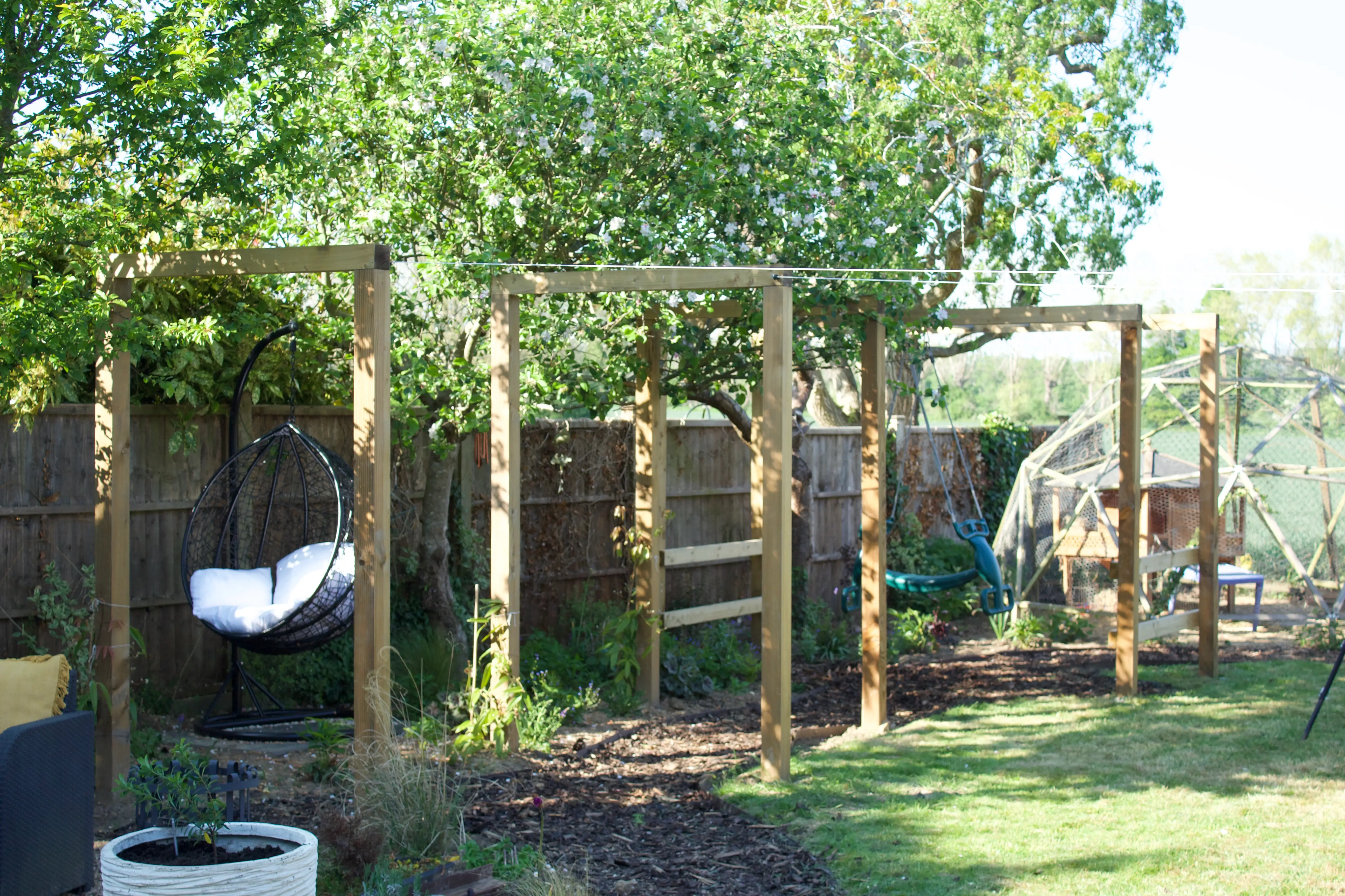 A wooden play structure in a grassy yard, surrounded by trees, with a slide and climbing features.