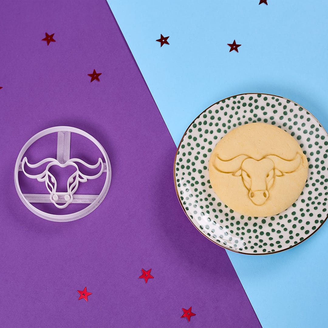 Okrągłe ciastko z wzorem znaku zodiaku Byka jest wyeksponowane na talerzu w kropki obok pasującej foremki do ciastek, na tle intensywnego fioletowego tła z rozsypanymi czerwonymi konfetti w kształcie gwiazdek, ukazując skomplikowane detale osiągalne dzięki foremkom do ciastek Biocraftlab.