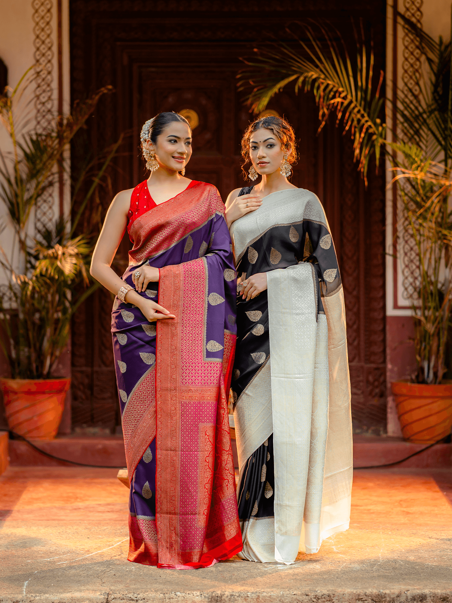 Two women standing together—one wearing a purple crepe silk saree with a pink-red contrast border and zari buttas, and the other wearing a black and white crepe saree—showcasing elegant handcrafted designs from That Saree Shop.