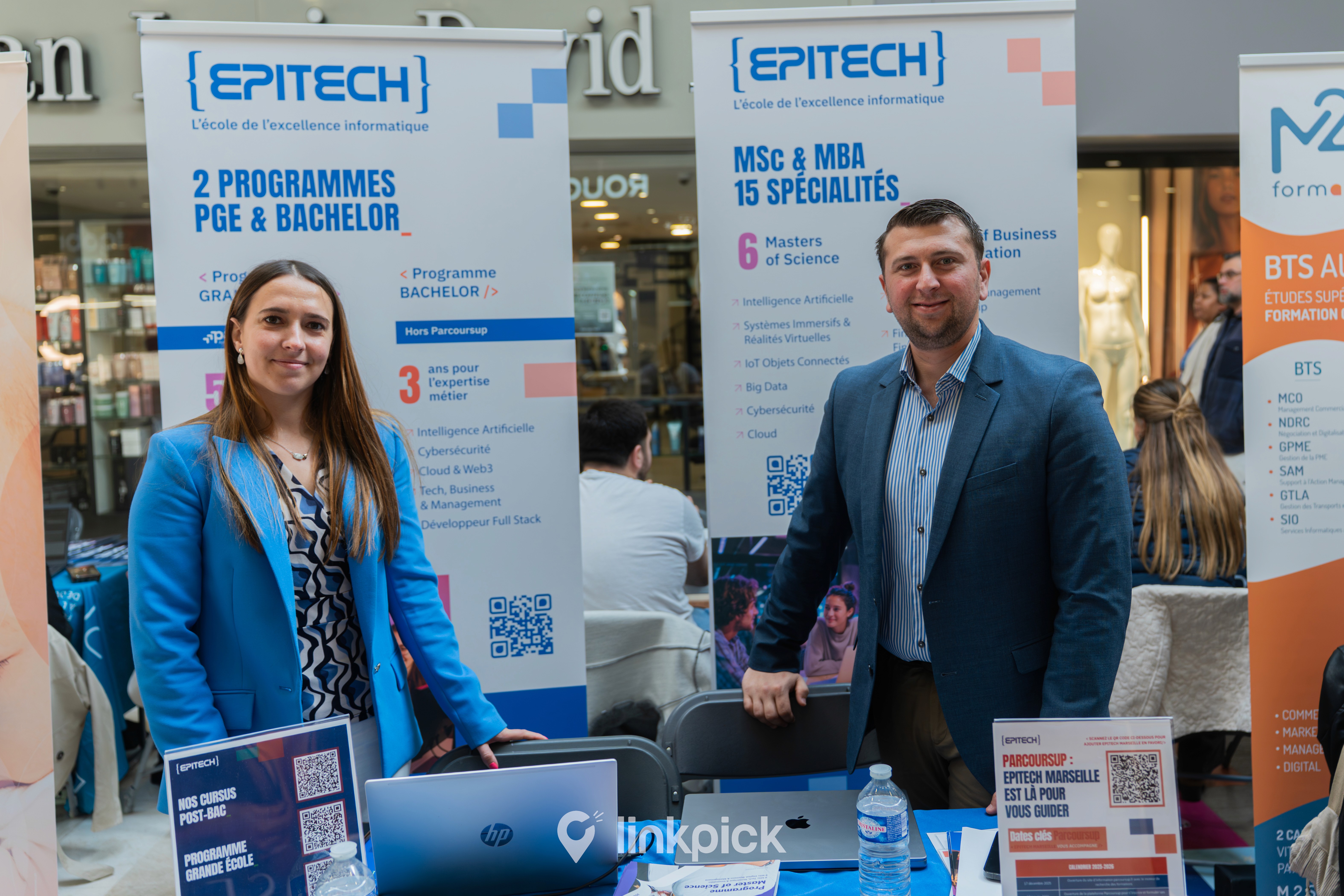 Marion Huc (Directrice adjointe d'Epitech) et Enzo Scaduto (Directeur d'Epitech) souriants derrière leur stand lors du Salon de l’Orientation et de l’Alternance. Ils sont entourés d'oriflammes présentant les programmes Grande École, Bachelor, MSc et MBA de l'école d'informatique Epitech. Le stand, installé dans les allées du centre commercial Grand Vitrolles, propose de la documentation et des QR codes pour guider les étudiants dans leur choix de formation.