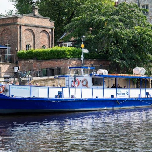 一艘名为“Witheycombe”的蓝白色游船停靠在一座砖砌建筑旁,附近有郁郁葱葱的绿树和石制结构。