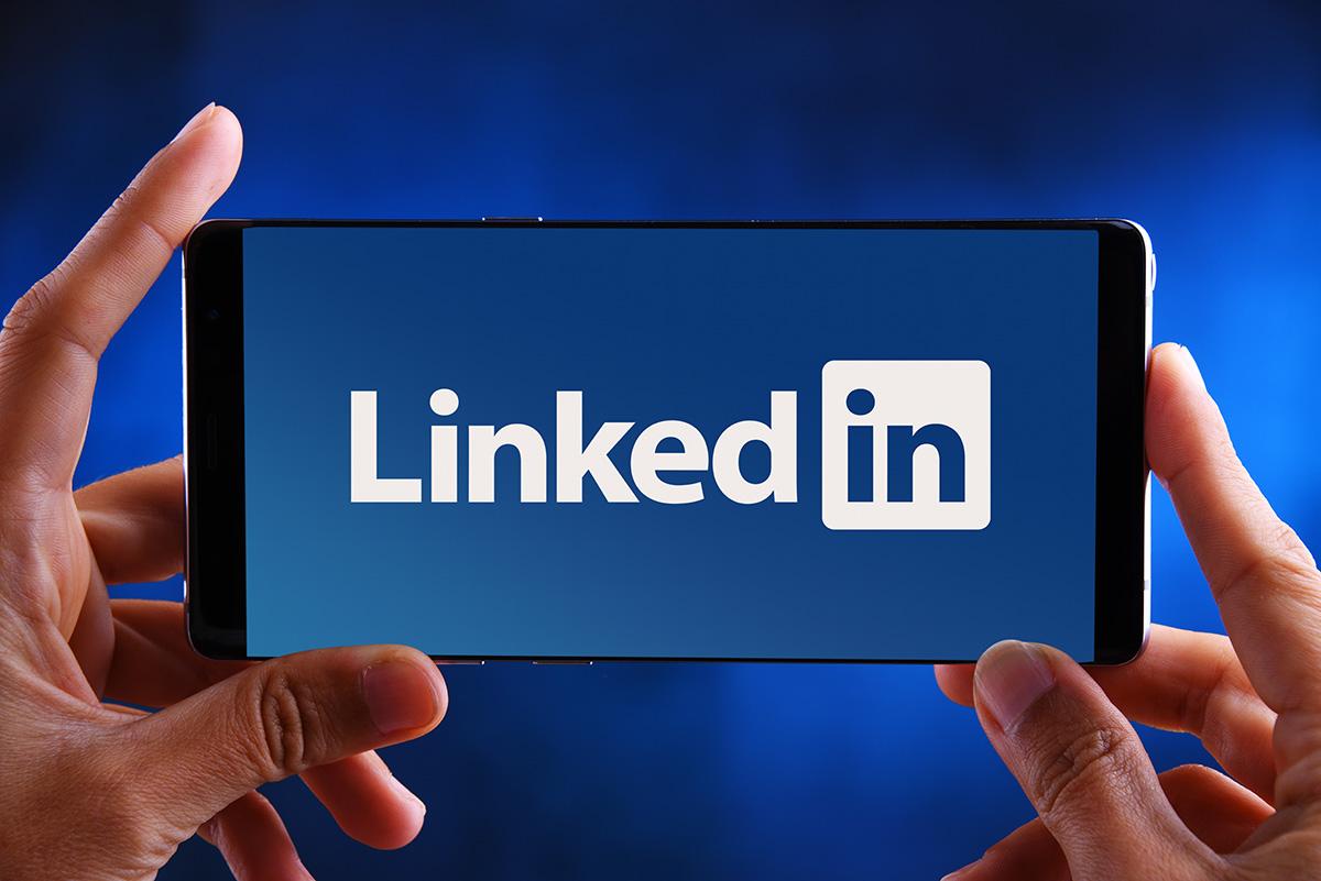 A hand visibly holding a phone tagged “LinkedIn” in bold letters. The image is placed on a gradient-blue background. 