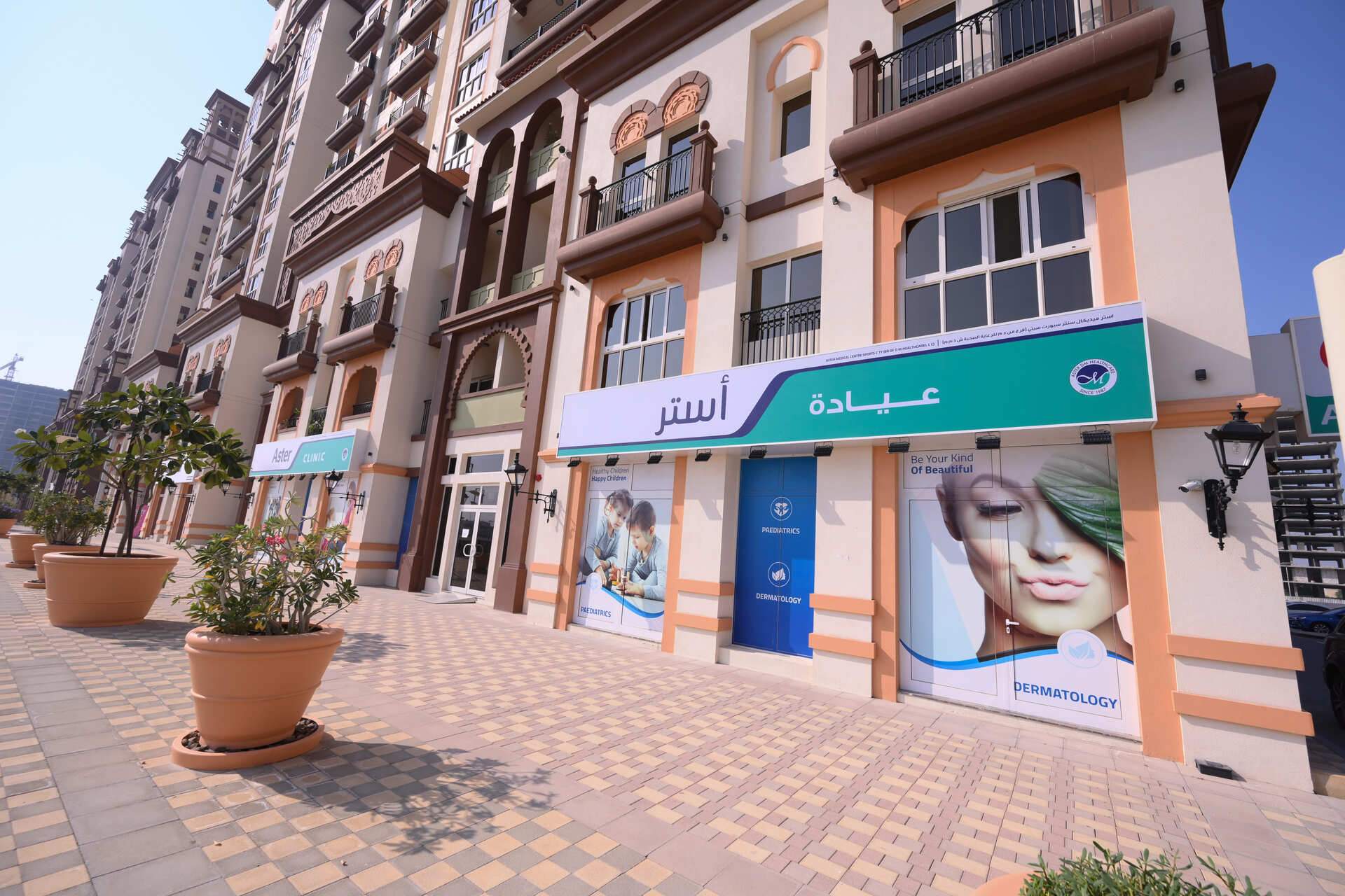 Aster Clinic at Dubai Sports City with several buildings close and tiled pedestrian walkway with potted plants.