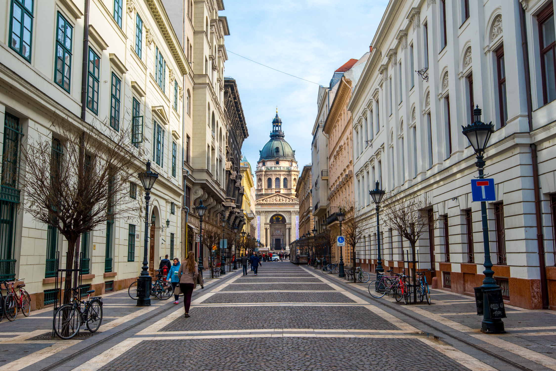 A long, paved pedestrian thoroughfare leads toward a grand, domed building under bright daylight, flanked by multi-story stone structures.