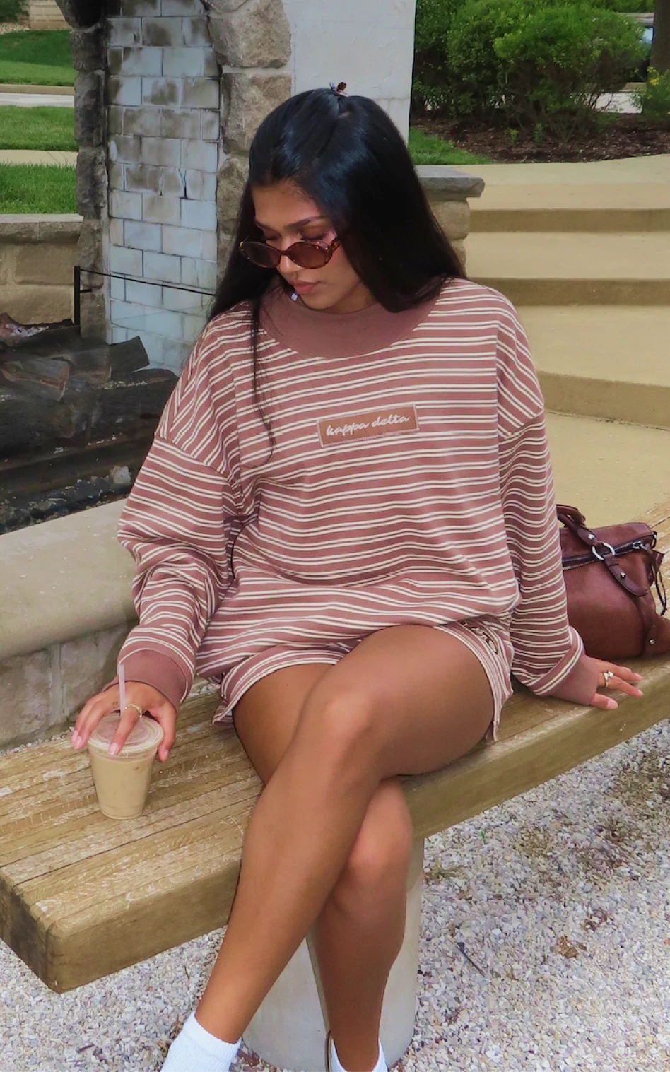 Sorority girl Meggie Eyer in a brown striped Kappa Delta sweatshirt, sunglasses on, sitting on a bench with an iced coffee.