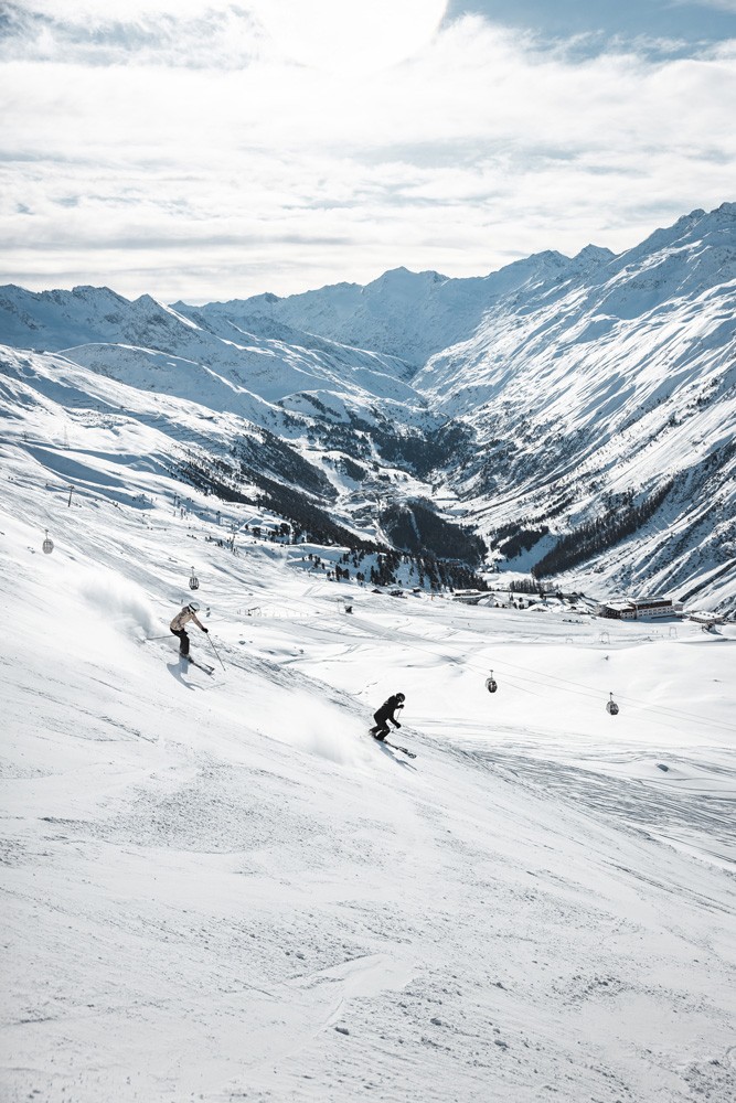 Zwei Skifahrer fahren einen schneebedeckten Berg im Ötztal hinunter, vor der atemberaubenden Kulisse zerklüfteter Gipfel unter einem teilweise bewölkten Himmel, umgeben von einer Reihe von Skiliftkabeln, die in der Ferne zu sehen sind.