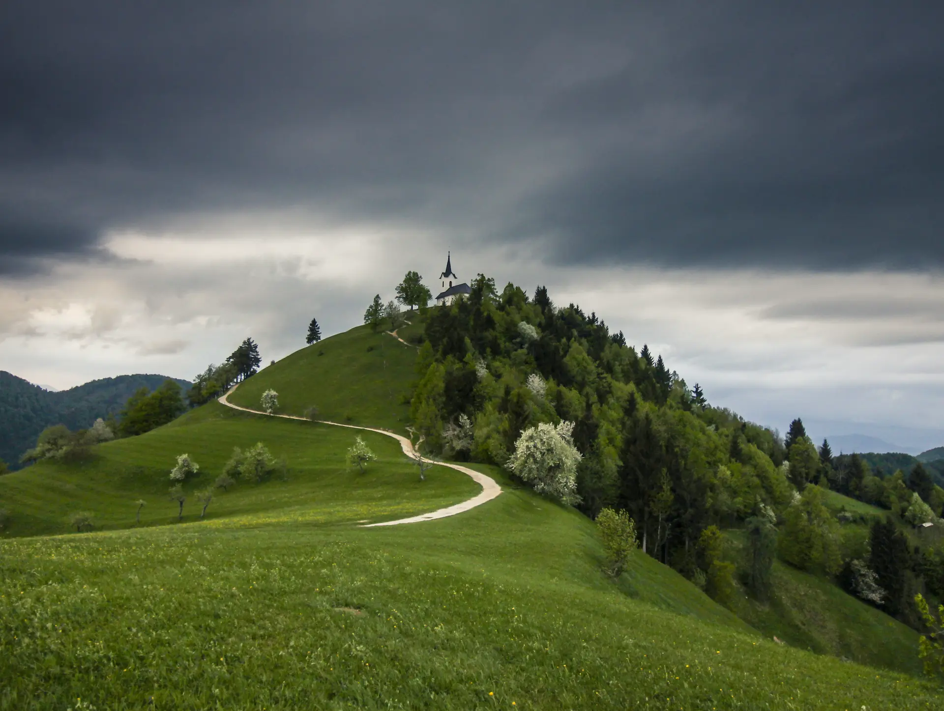 Sveti Jakob church in Slovenia on a grassy hilltop under dark stormy skies.