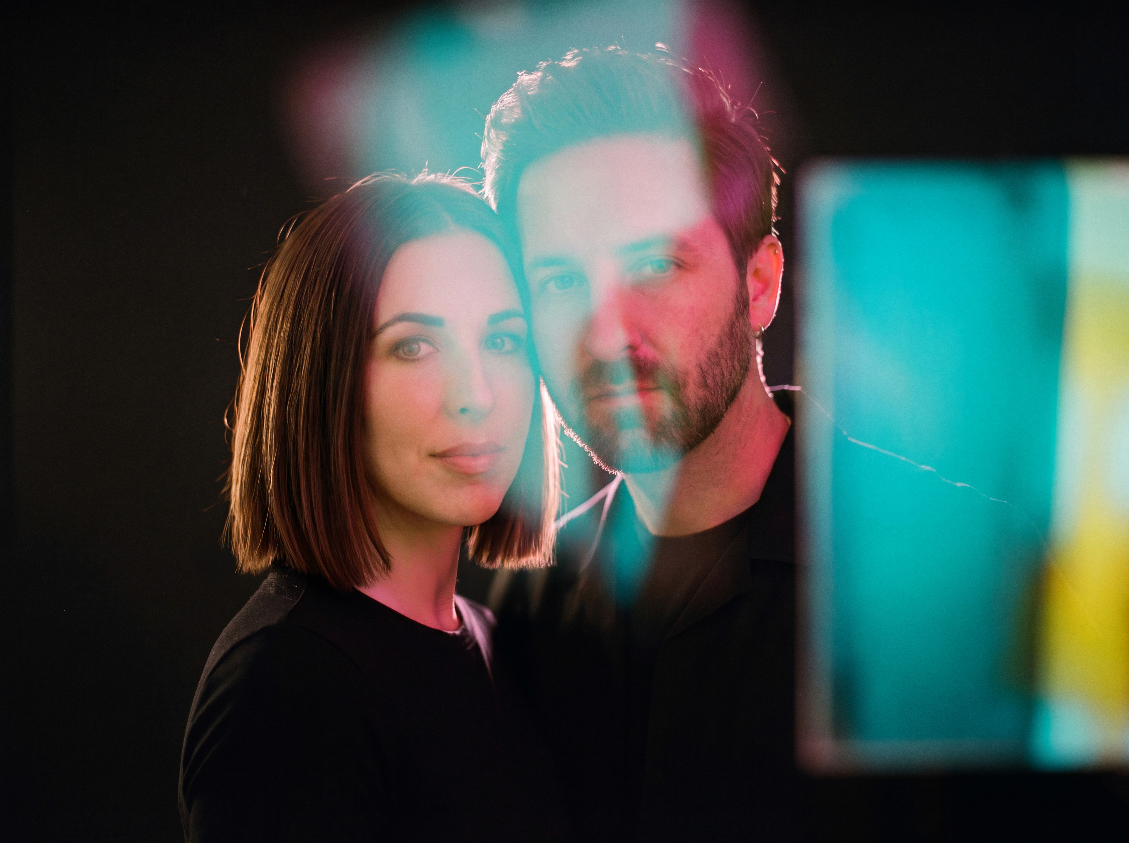Kyle and Rachel Ford in 2026, photographed on film through a sheet of glass carrying washes of teal and magenta light, ten years after founding Ford Media Lab.