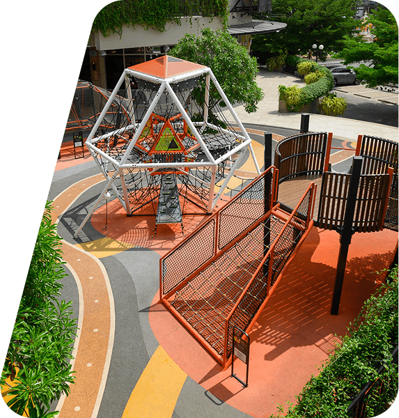 Colorful playground with intricate climbing structures, including a netted white and orange dome, set on a bright, winding path. Lush greenery surrounds the area, giving it an inviting and vibrant atmosphere.