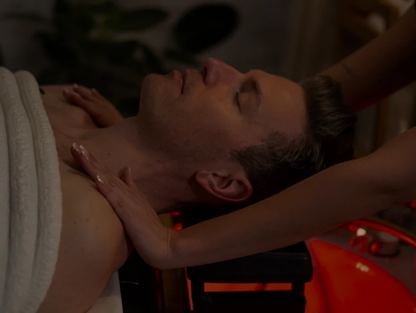 A person lying comfortably under a white towel receives a relaxing neck and shoulder massage in a dimly lit spa, enhancing a serene and rejuvenating atmosphere.