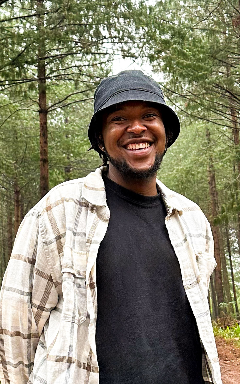 A medium shot of Ashley Ncube, a UI/UX Designer from South Africa, standing tall amongst the green trees, whilst it rains