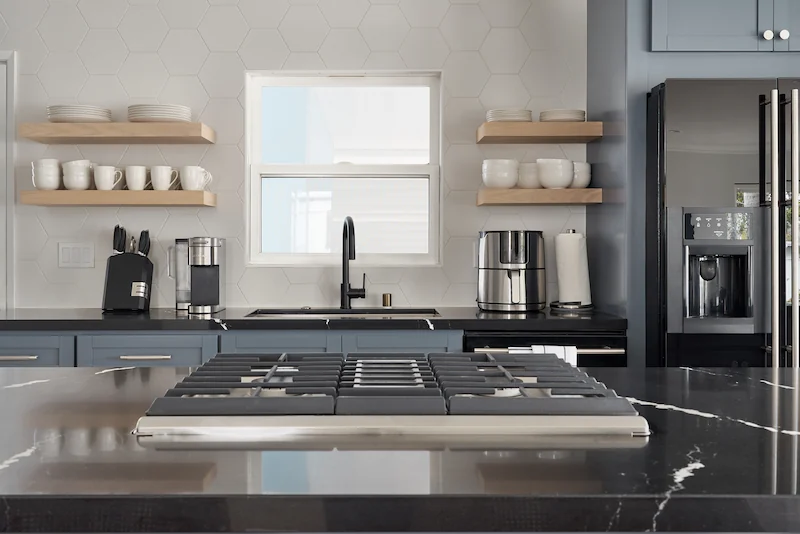 Modern kitchen with gray cabinets, stainless steel appliances, floating shelves, and a central cooktop island. Sunlight filters through a white-framed window. Photo by Todd Huge.