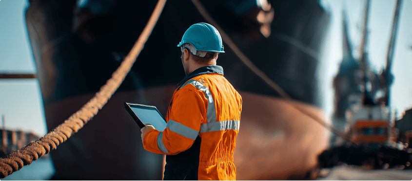 A crew member in an orange suit approaches a large cargo ship while others climb the side of the vessel.