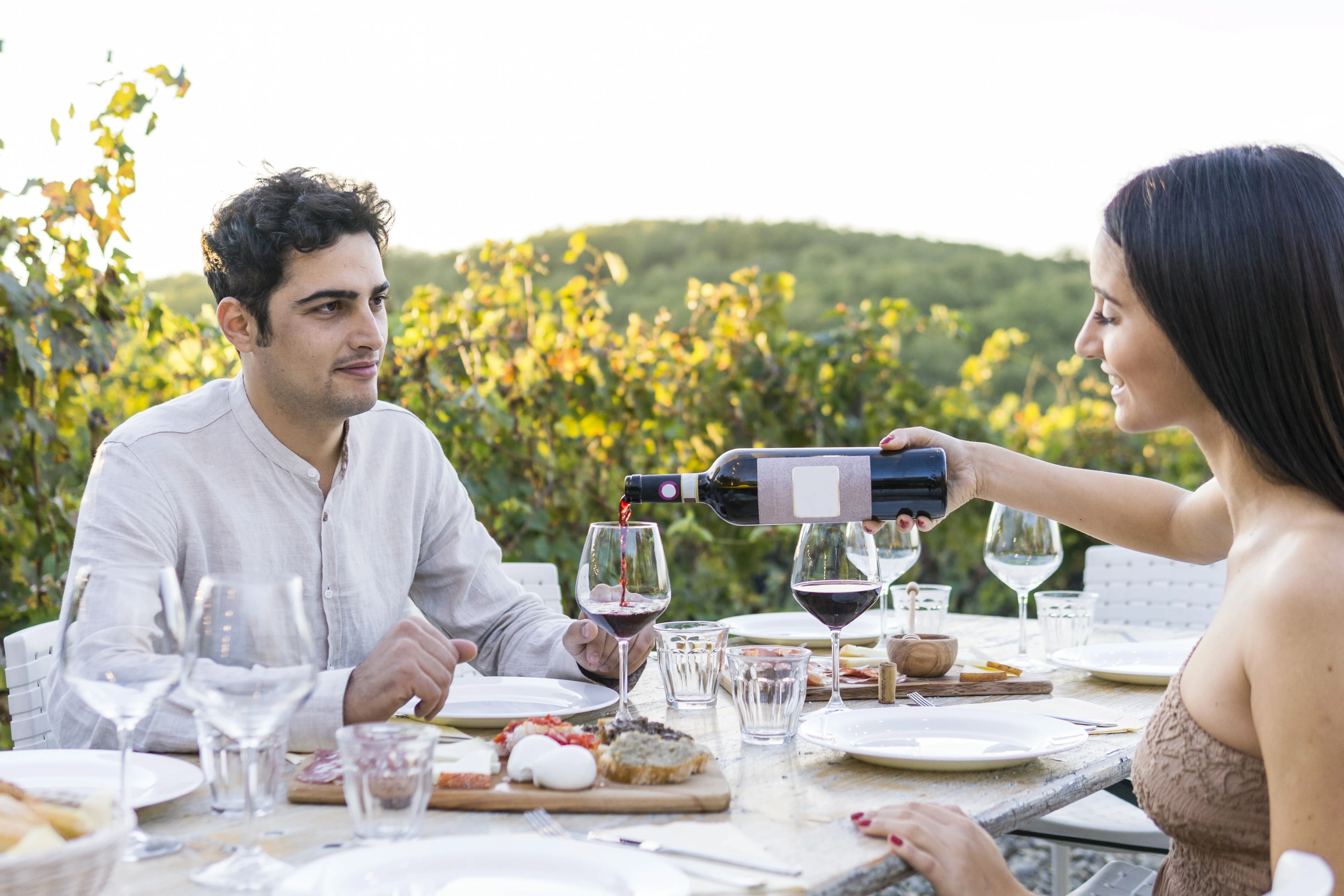 A woman pouring red wine for a man during an outdoor meal at a vineyard.