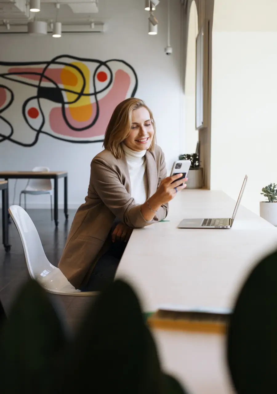 Photograph of a woman smiling, sitting at a desk in front of a laptop, holding a smartphone in one hand