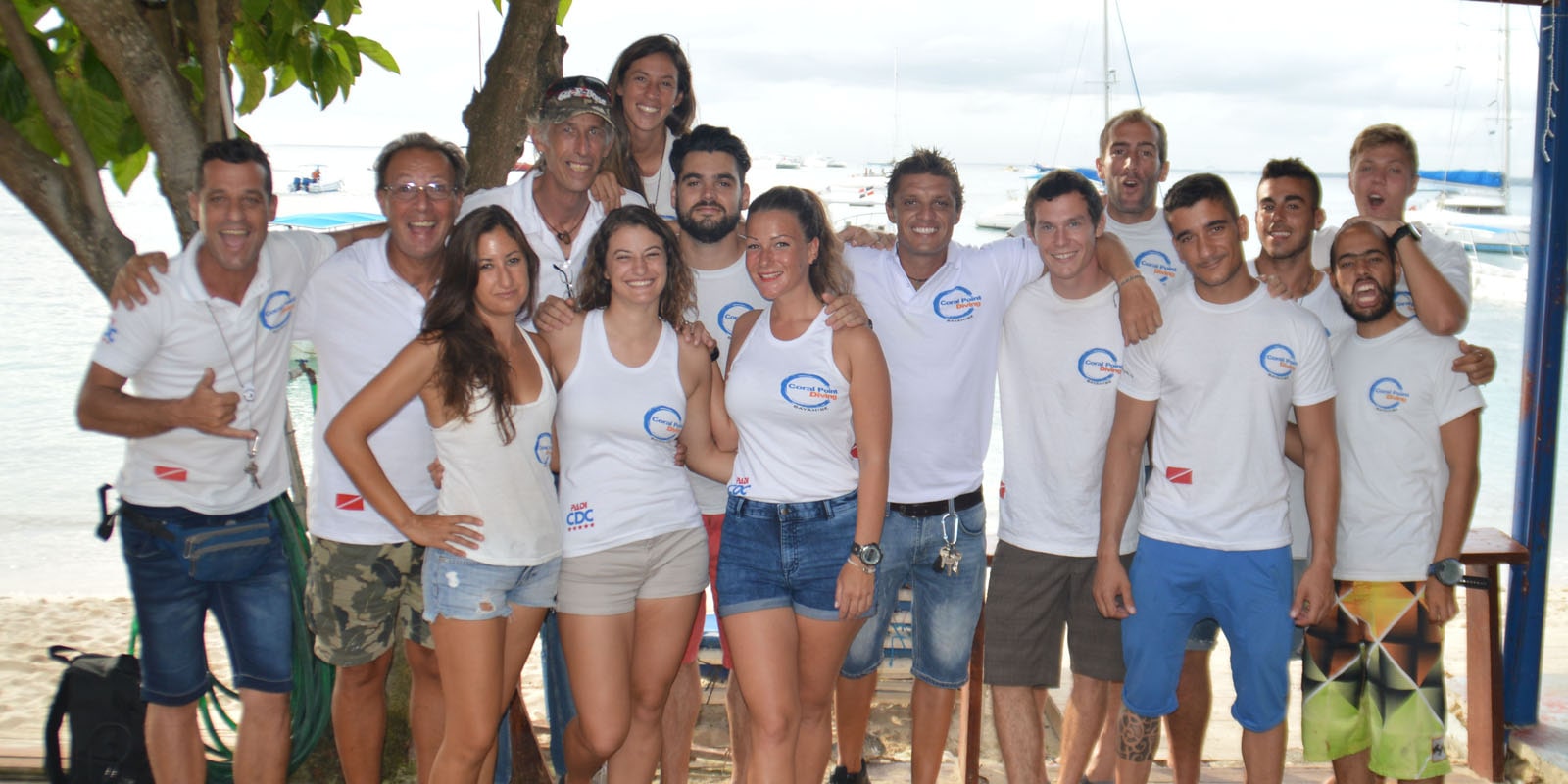 Group of smiling adults wearing matching white shirts posing together on a beach, with boats and water visible in the background.