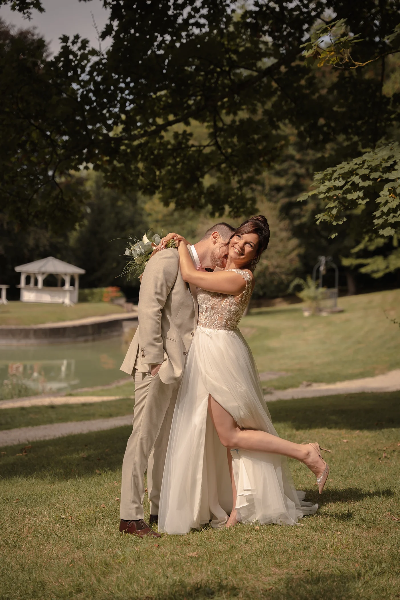 Smiling bride kicking up her heel while hugging the groom in a beige suit, in a park with a pond and white gazebo