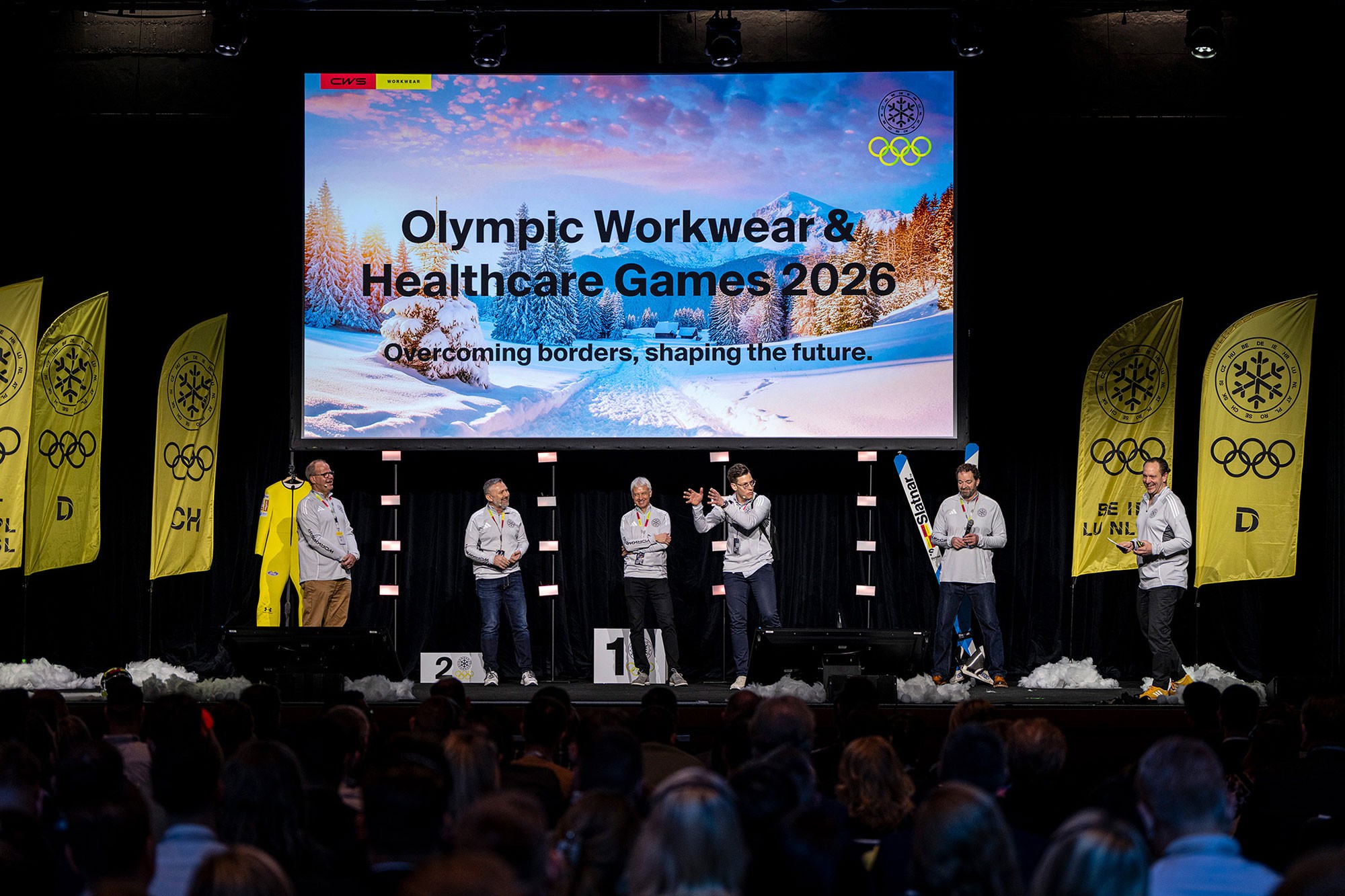 Speakers on stage at the CWS Olympic Workwear & Healthcare Games 2026 sales conference in Prague, presenting in front of a large branded screen and audience.