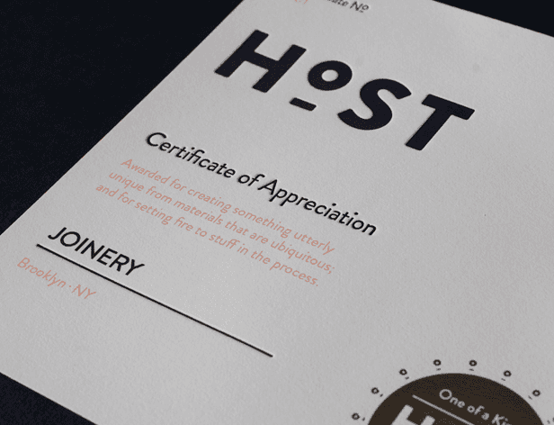 A white certificate titled “HOST” with the subtitle “Certificate of Appreciation” printed below. The certificate is signed “JOINERY” in black ink, with “Brooklyn, NY” written below it in orange ink. A small circular design with “One of a Kind” is visible in the bottom right corner.
