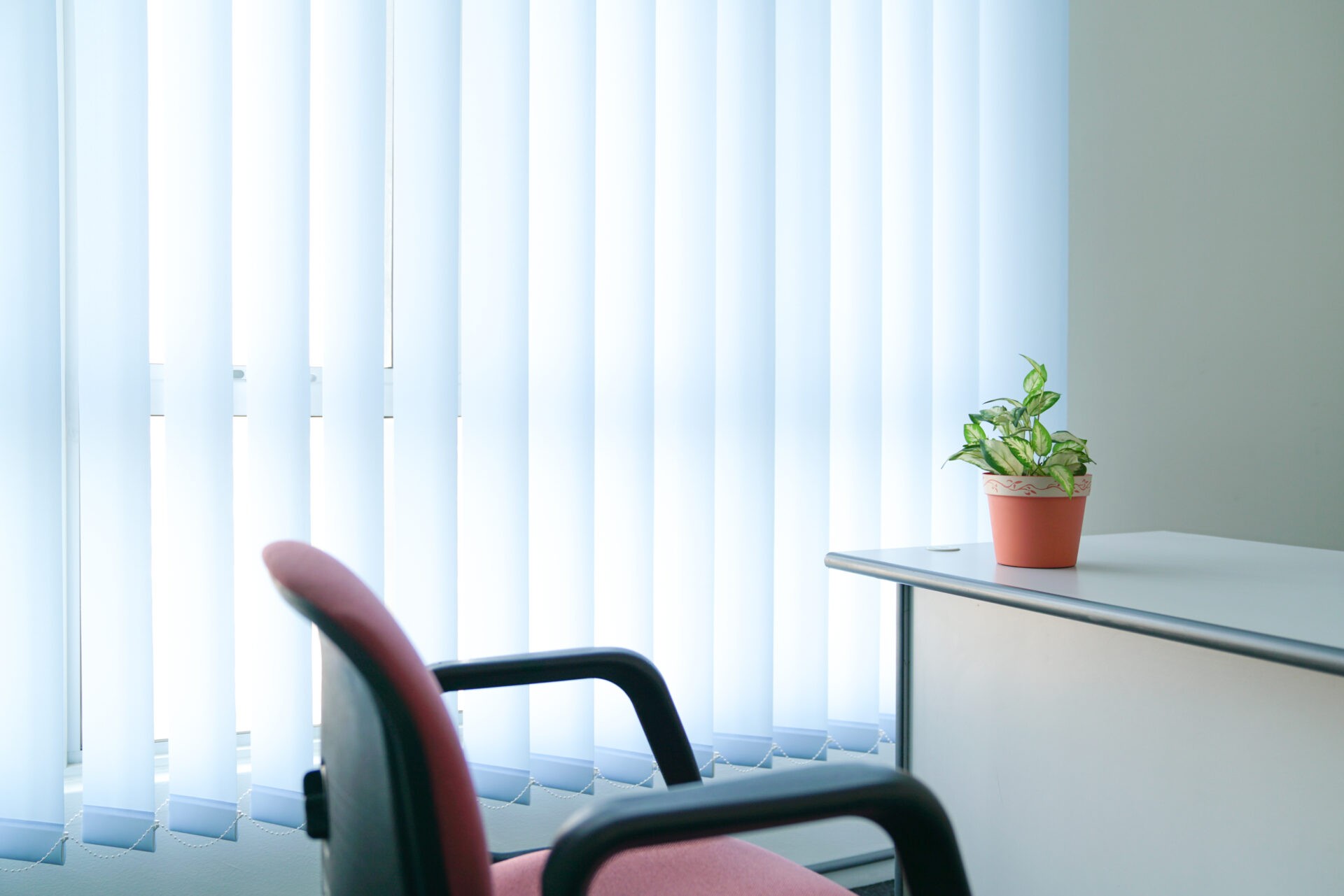 Vertical blinds in a bright office, red office chair and office plant. Wholesale Blind Factory