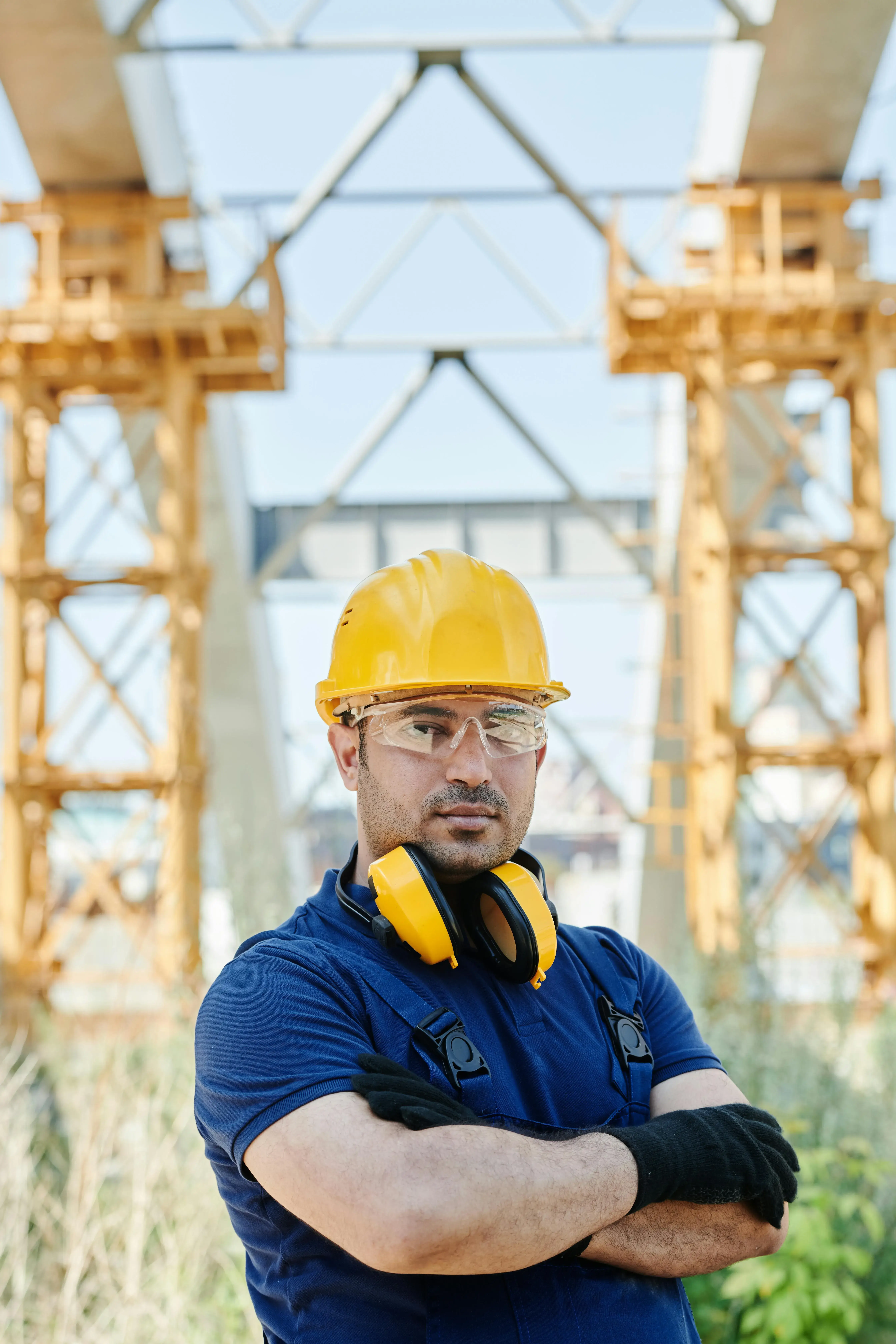 Construction worker in a blue shirt with a yellow hard hat, safety glasses, and headphones around his neck stands confidently, crossed arms, amidst metal scaffolding.