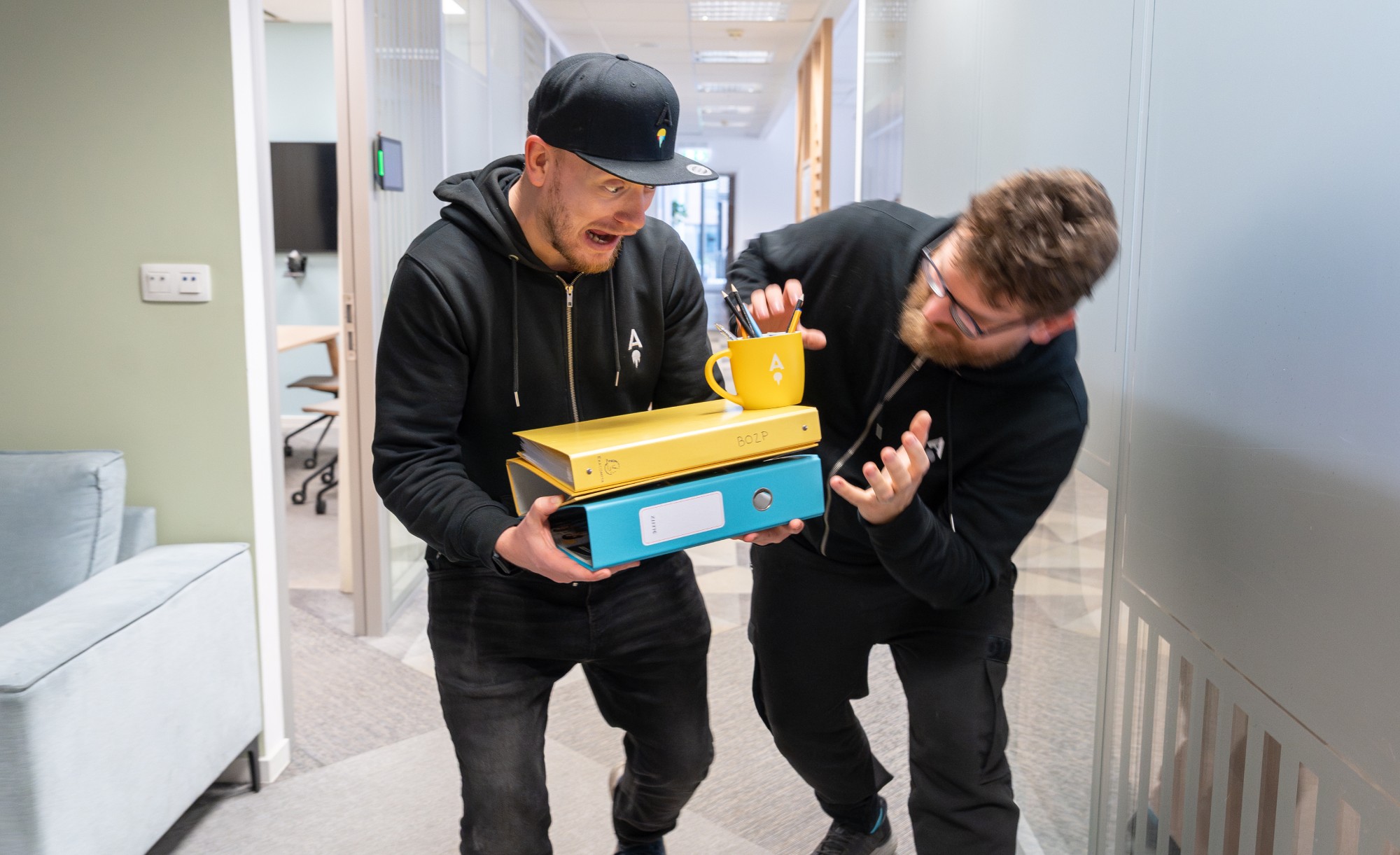 Vratislav and Filip humorously struggling to carry a stack of colorful office binders and a mug.