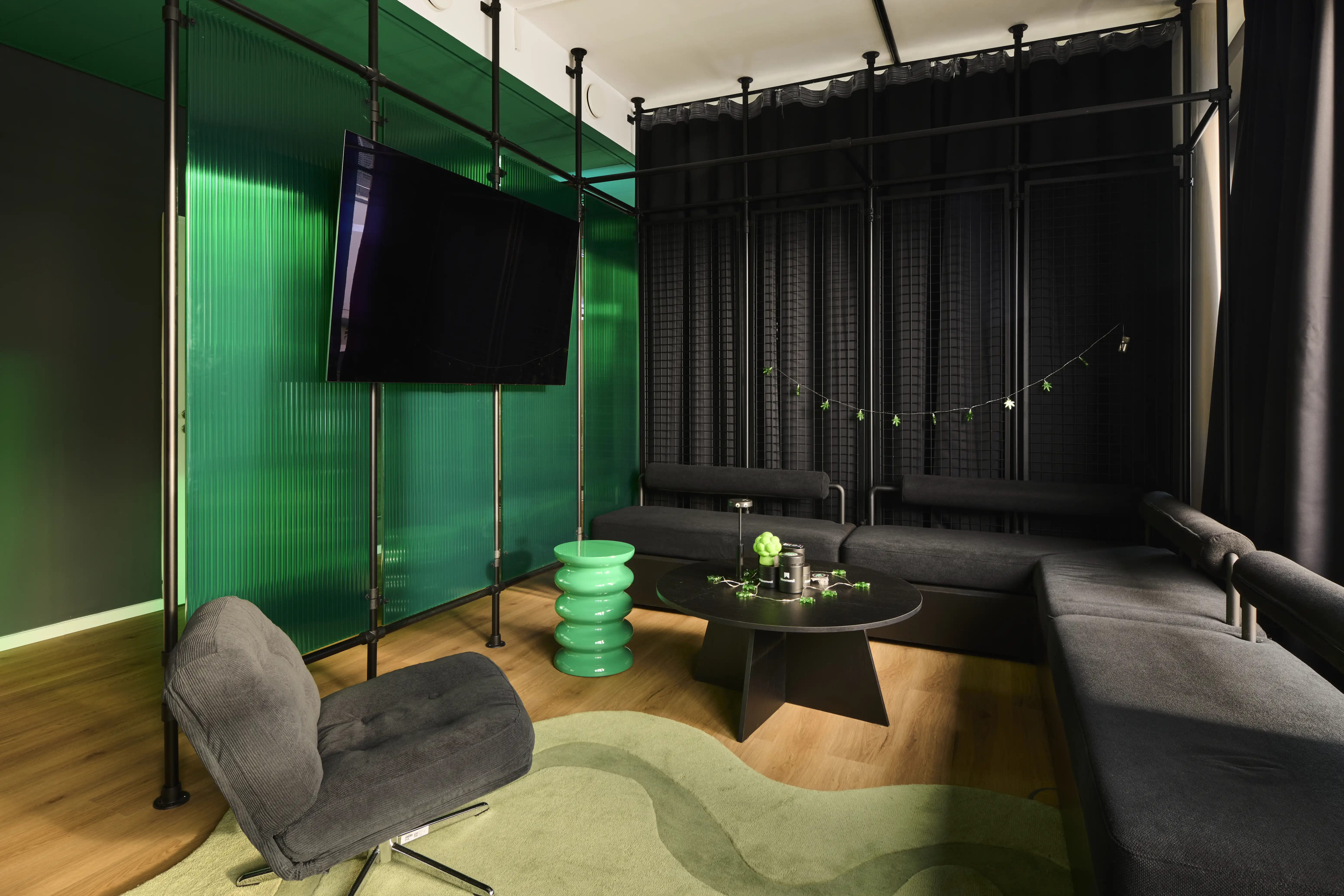 Lounge at BLOOMWELL with charcoal sofas around a black round table, a mint-green ribbed side table, a TV on a black steel frame against a green glass wall and a wavy green rug.