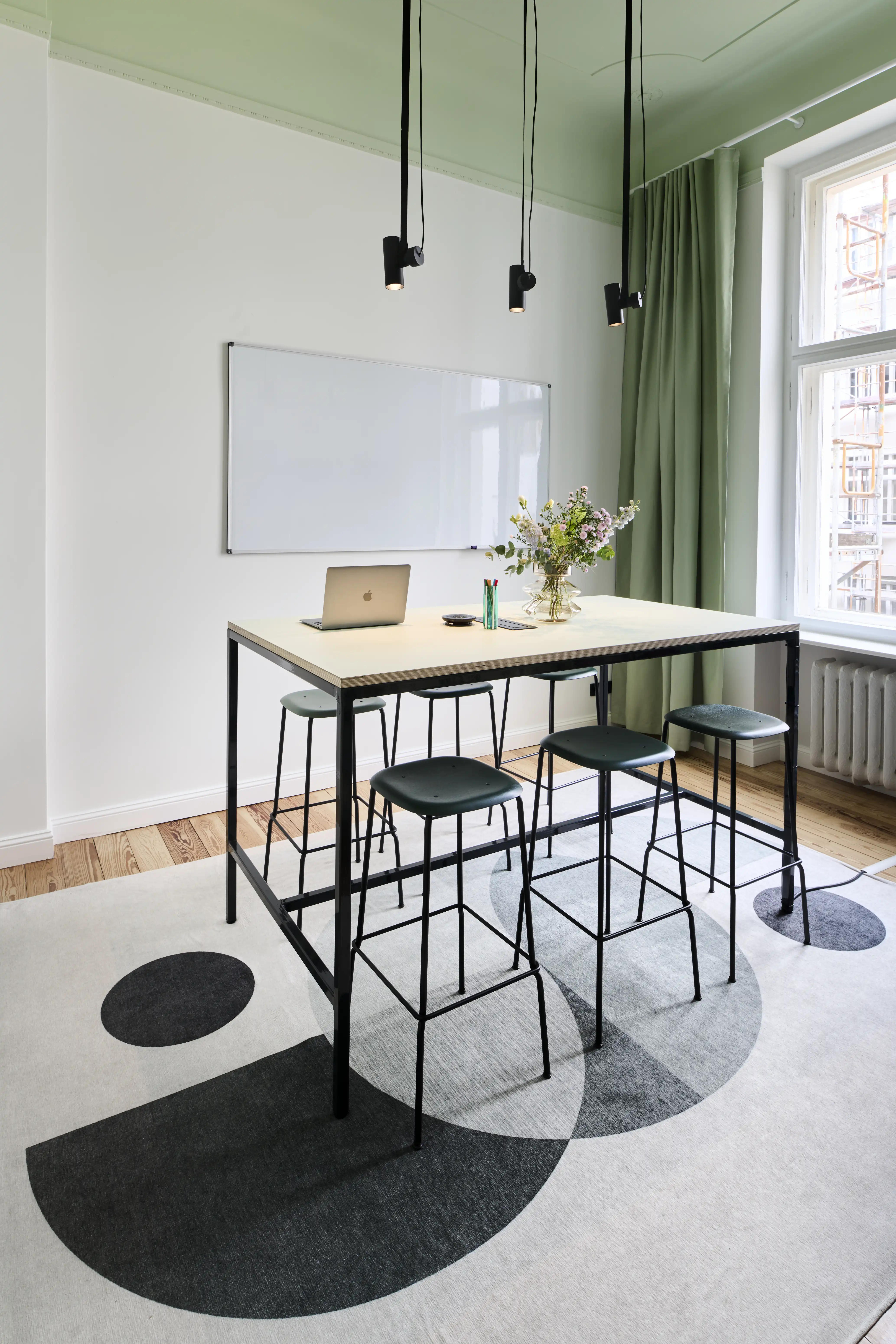 Standing meeting room at INTERMATE BERLIN with a black-framed high table, dark green bar stools, a whiteboard, sage curtains, green ceiling and a rug with black circle pattern.