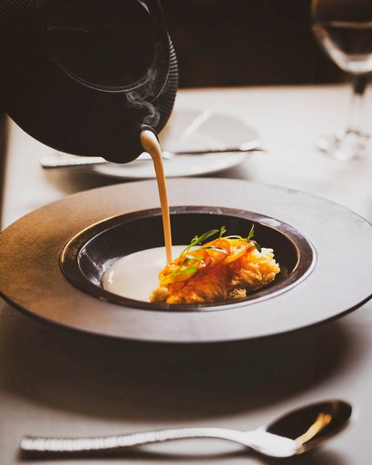 Creamy soup being poured into a bowl with grilled fish and garnish.