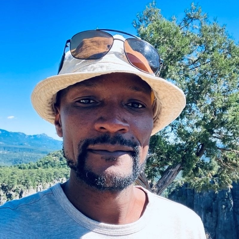 A man wearing a white bucket hat with sunglasses perched on top faces the camera, he has a short dark beard, in the background are blue skies, trees and mountains