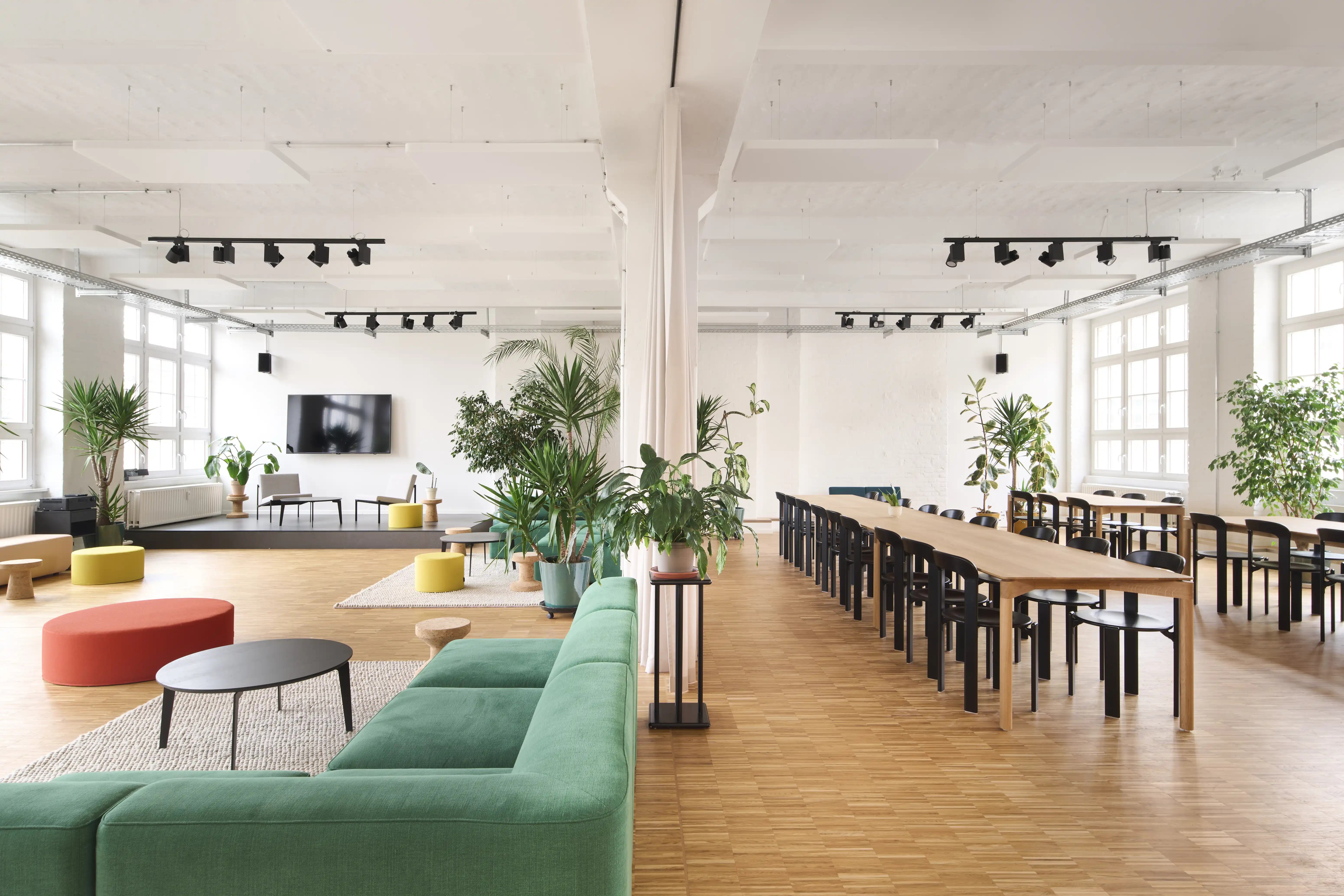 Loft office at PLAN A with a green modular sofa and colourful poufs on the left, long oak dining tables with black wooden chairs on the right and white beamed ceilings with black spotlights.