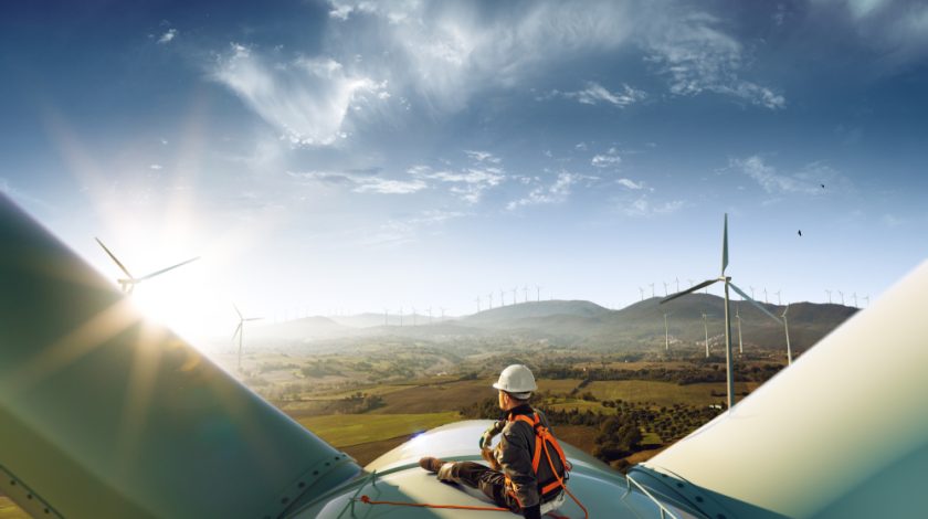 A person in safety gear sits atop a wind turbine, overlooking a vast landscape of rolling hills dotted with several other wind turbines under a clear blue sky, capturing the essence of renewable energy and sustainable technology.