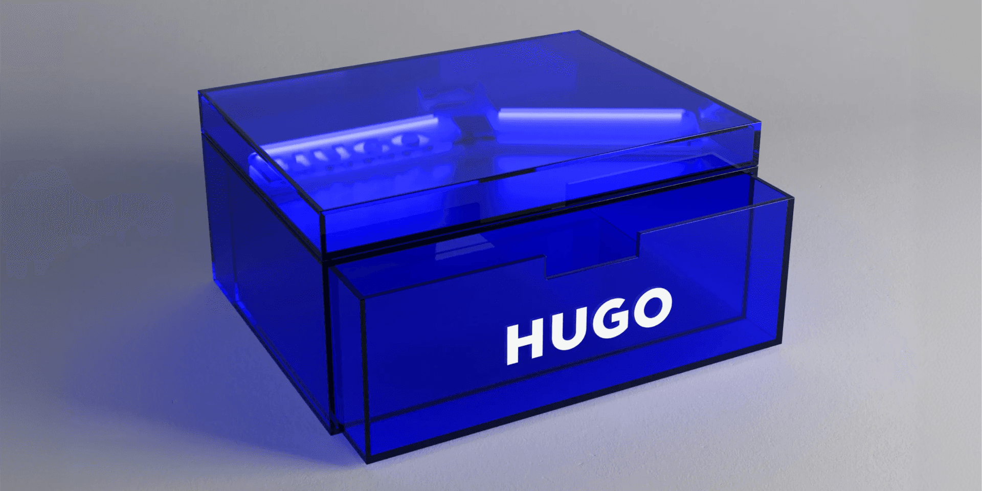 Markenwelt HUGO – transparente Acrylglas-Box mit Schubfach und Deckel in Hugo-Blau, weisses „HUGO"-Logo frontal, vor neutralem Hintergrund inszeniert.